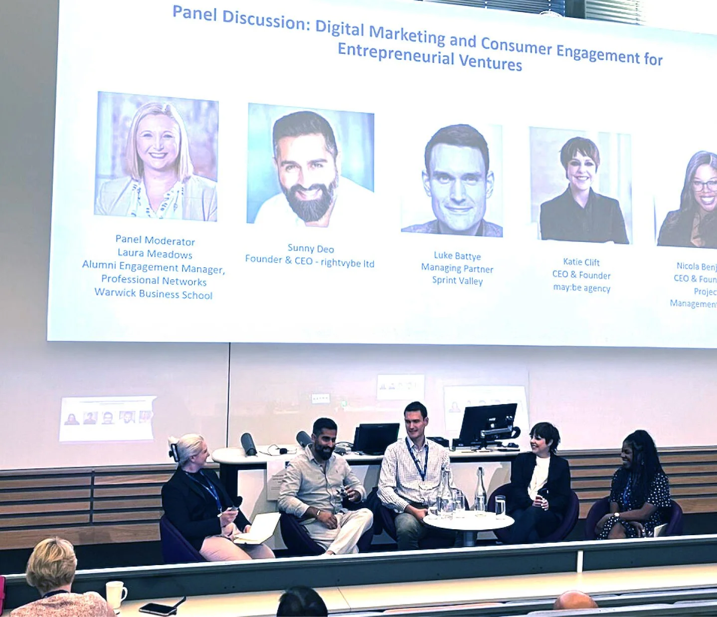 Panel discussion on digital marketing and consumer engagement for entrepreneurial ventures with five panelists sitting on stage. A large screen behind them displays profile photos and names of the panelists along with their titles and affiliations.