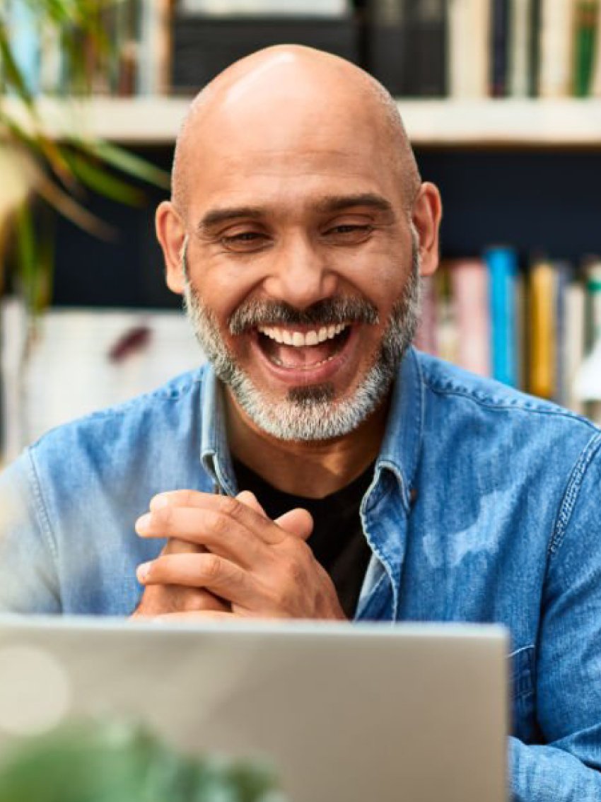 A middle-aged man with a shaved head and gray beard, laughing while looking at a screen, wearing a denim shirt, in a room with a bookshelf in the background.