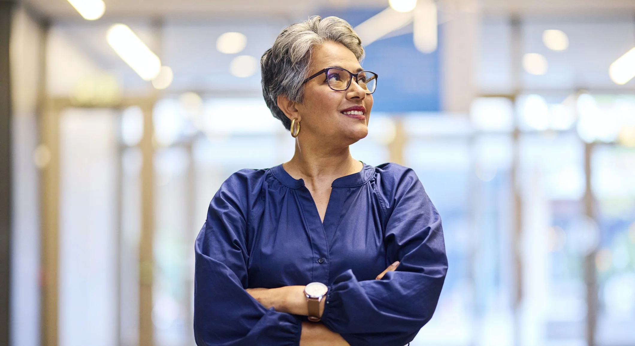 A confident senior woman with short gray hair, glasses, and gold hoop earrings, standing with arms crossed in a modern, well-lit indoor space, wearing a navy blue blouse and a watch.