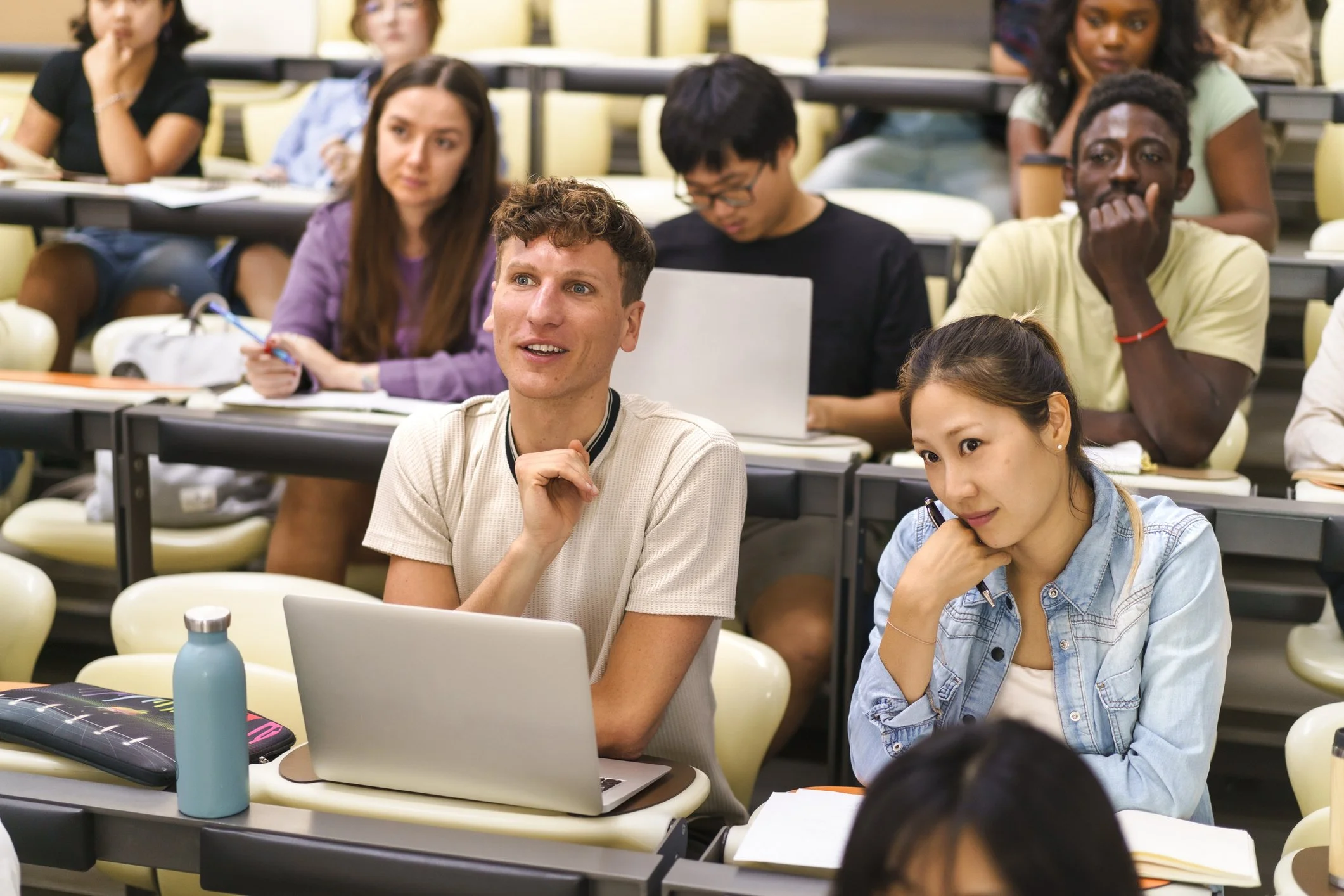 A diverse group of students in a classroom, some engaging with laptops and notebooks, listening attentively.