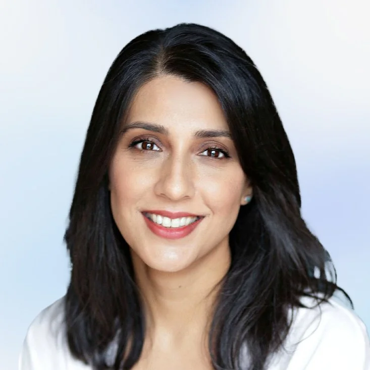 A woman with long black hair, light skin, and a bright smile, wearing a white top, standing against a light blue background.