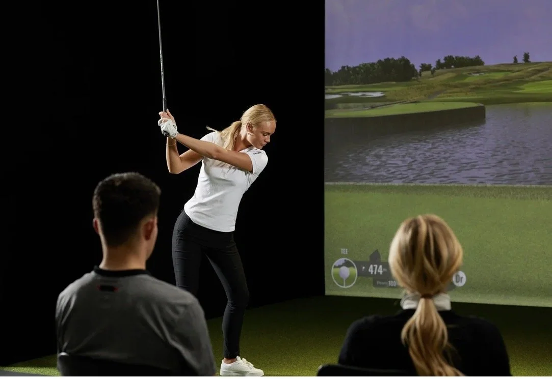 A woman is practicing golf indoors using a simulator, with two people watching.