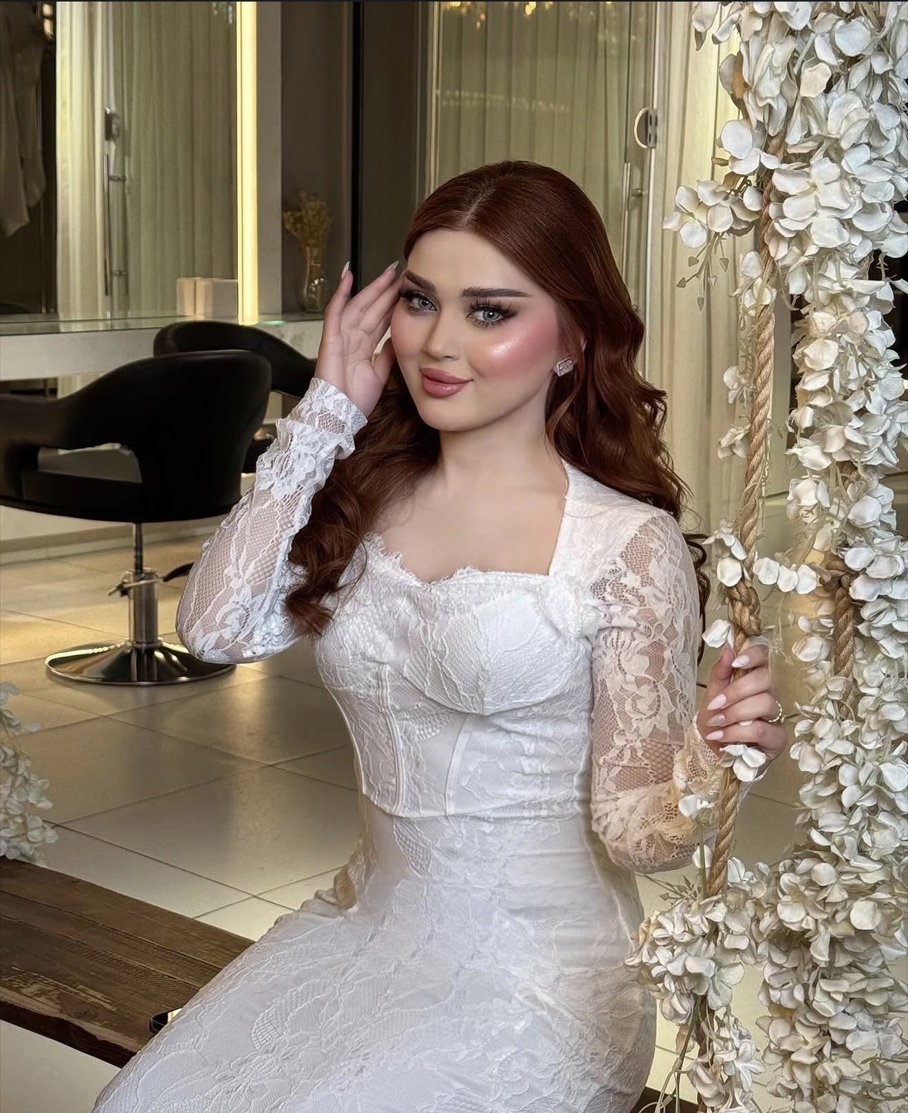 A woman with long red hair and makeup, wearing a white lace dress, is seated near a swing decorated with white flowers, in an indoor setting.