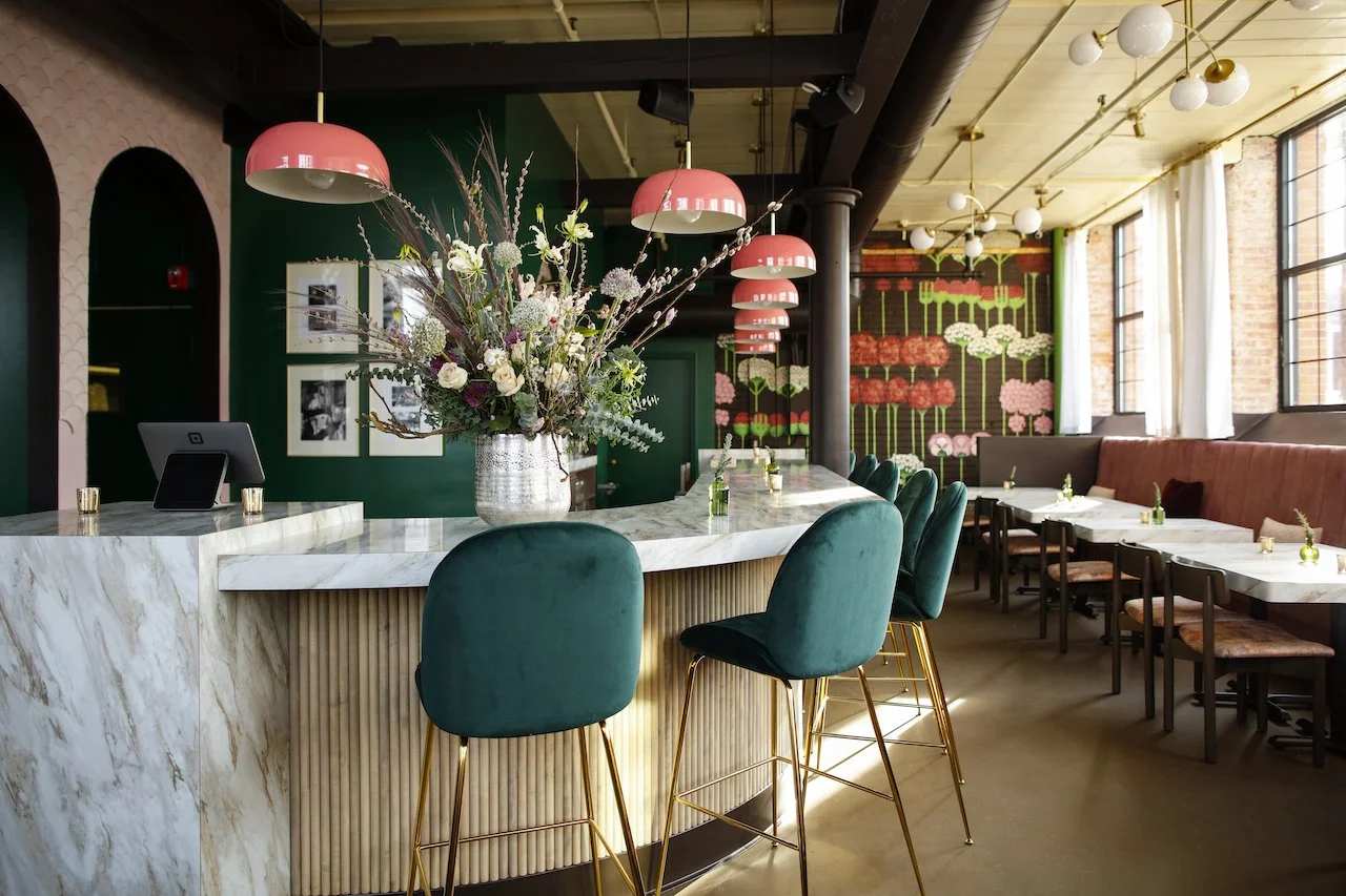 Interior of a modern restaurant or cafe with a marble counter, green velvet bar stools, and a large floral arrangement. The space has pink hanging pendant lights, natural light from large windows, and a decorative floral mural on the back wall.