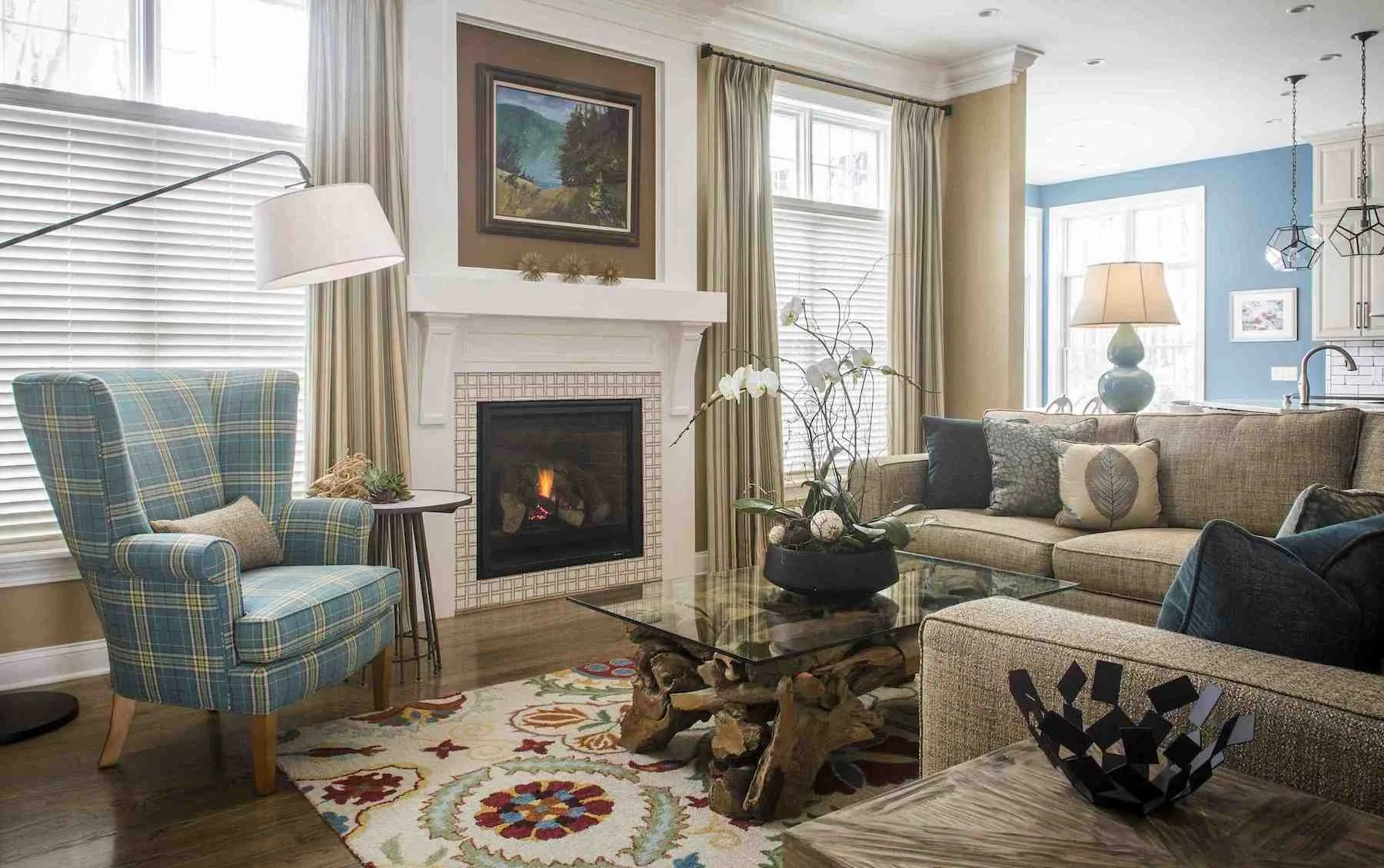 Living room with fireplace, plaid armchair, glass-top coffee table with wooden base, beige sofa with decorative pillows, window with blinds and curtains, and various lamps and artwork.