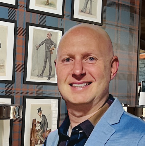 A smiling man in a light blue blazer standing in front of a wall decorated with framed illustrations of classical musicians.