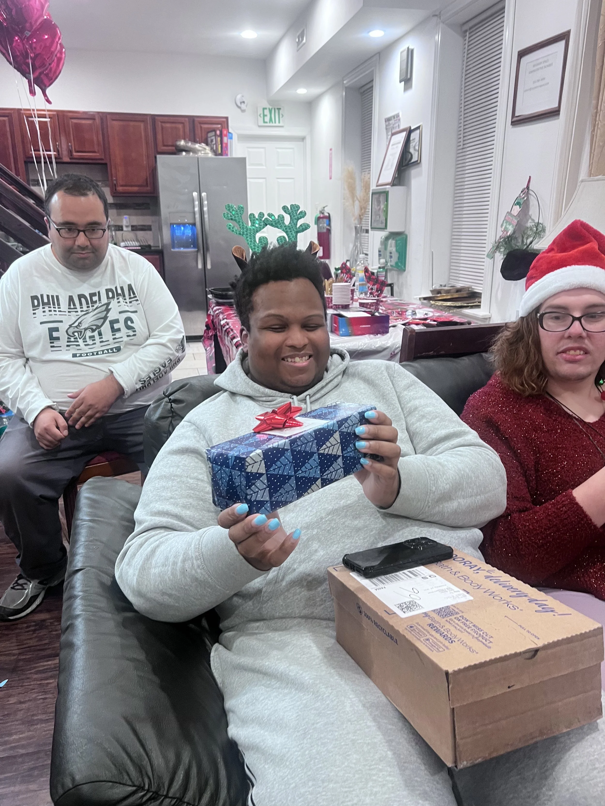 Three people at a festive gathering, one sitting on a couch holding a wrapped gift with a red bow, another sitting on the right wearing a Santa hat and glasses, and the third person standing in the background with reindeer antlers headband, in a room decorated for Christmas with balloons, a table with food and drinks, and holiday decorations.