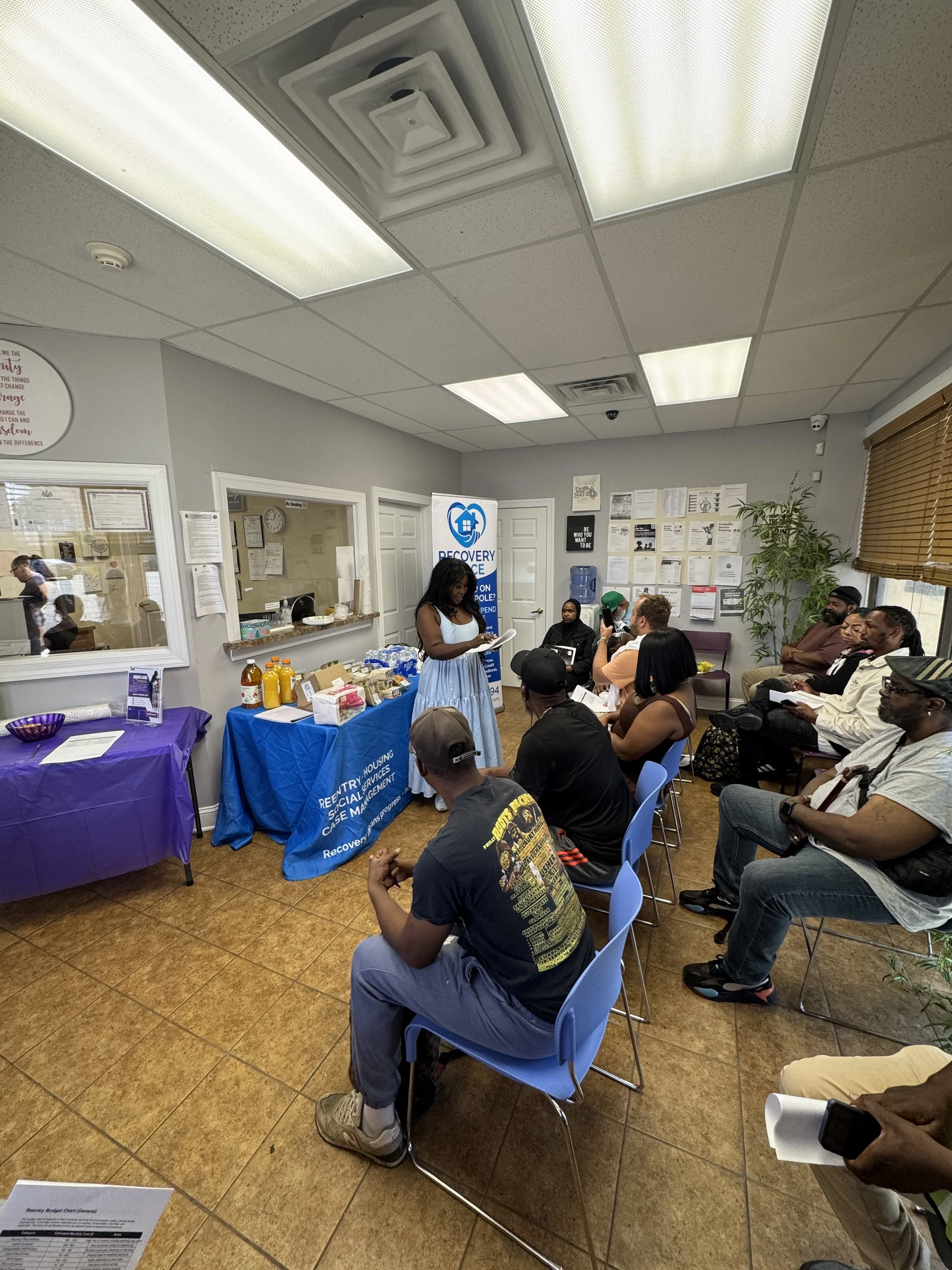 A group of people attending a presentation or workshop in a small room, sitting on blue chairs, with a woman speaking at a table with informational materials and a banner behind her.
