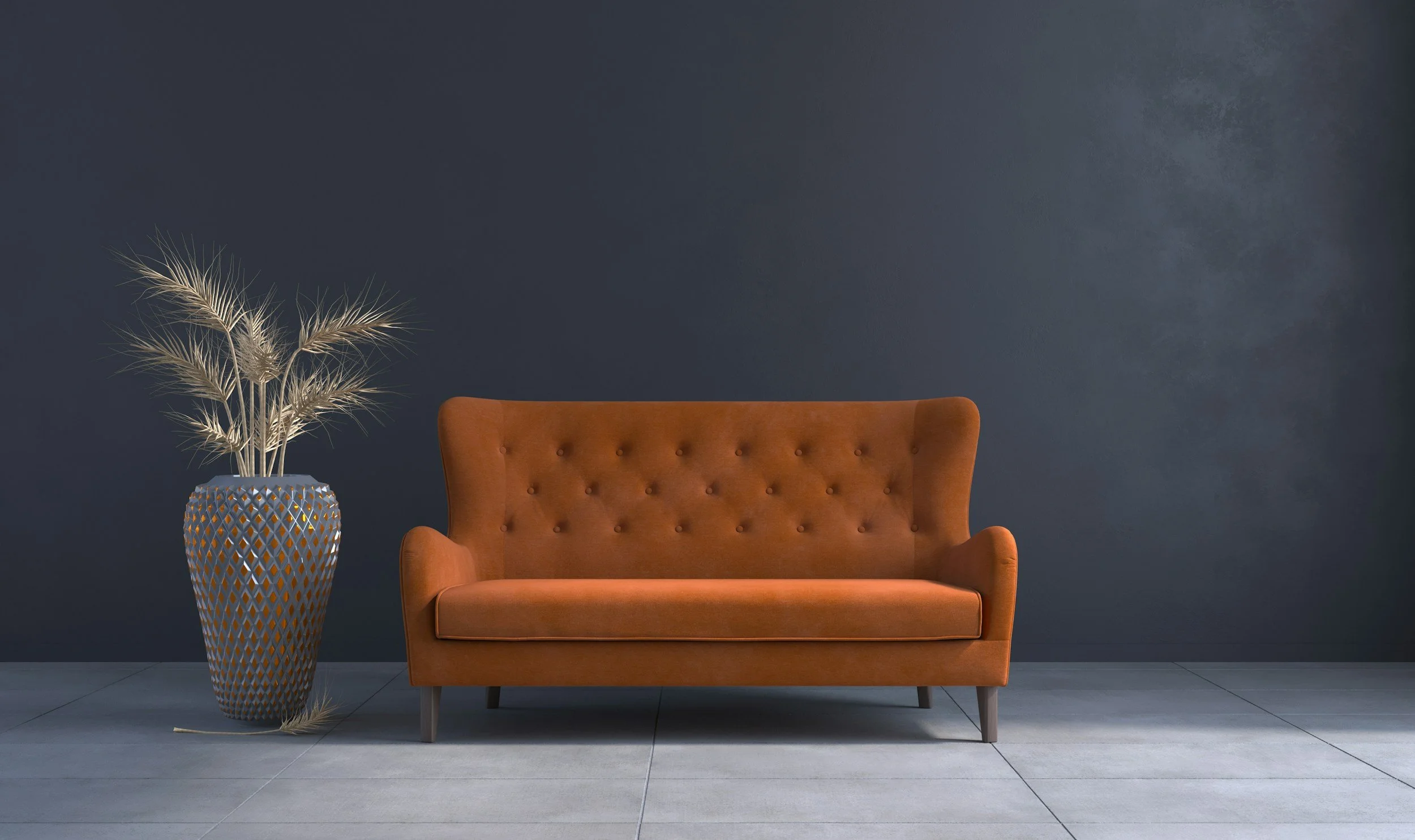 A burnt orange velvet tufted loveseat with wooden legs positioned against a dark wall, with a tall decorative vase containing dried beige palm leaves to the left.