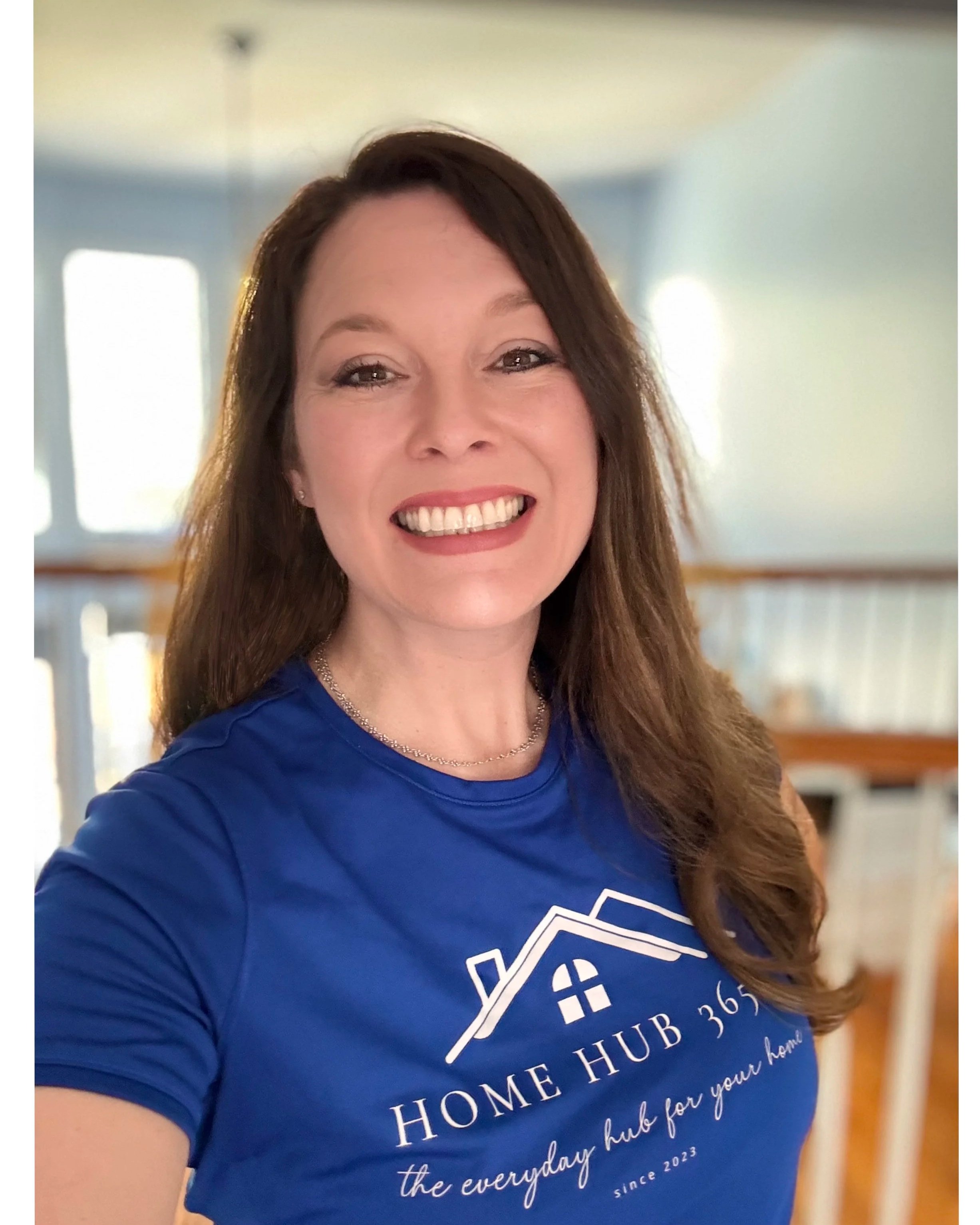 Tiffany Tanner Wilson smiling indoors in a home. She is wearing a blue T-shirt with a house logo and text that reads 'Home Hub 365, the everyday hub for your home, since 2023.' She has long brown hair and is wearing a necklace.