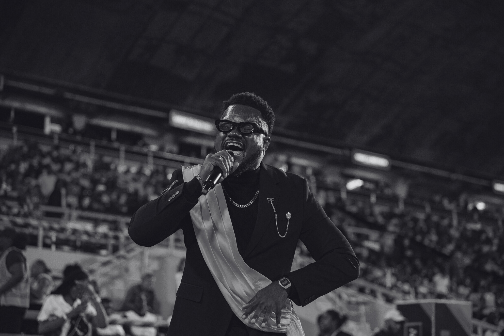 A male performer passionately singing into a microphone at an indoor venue, with an audience in the background.