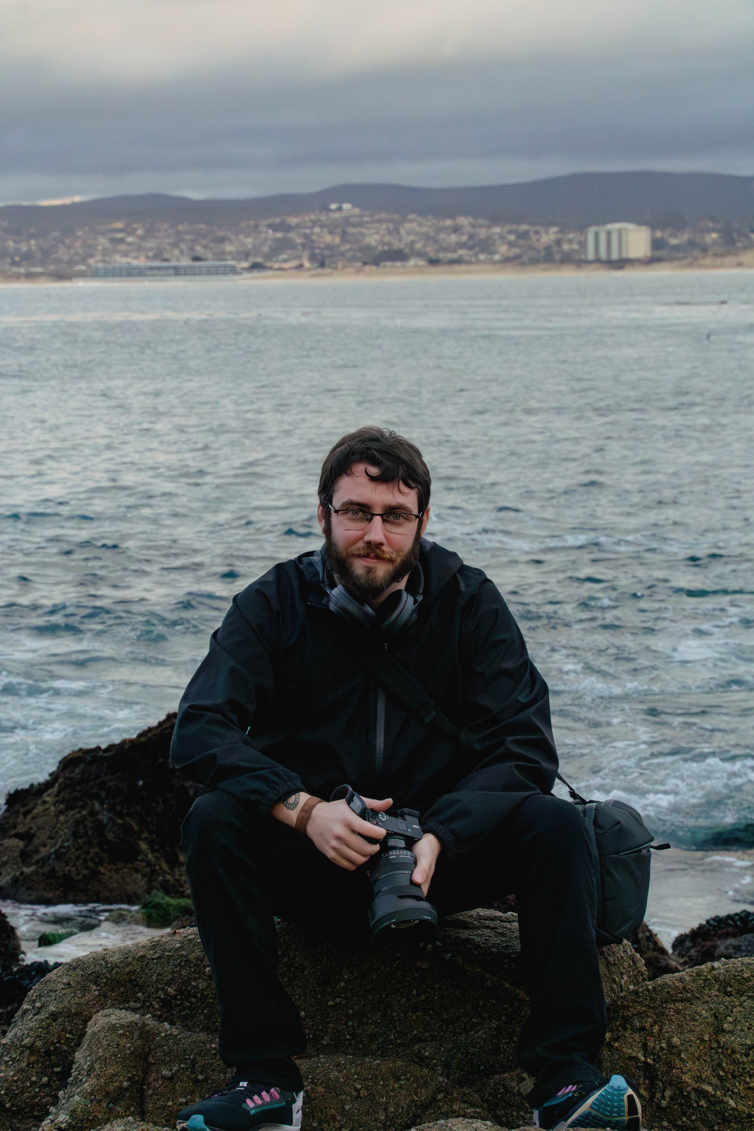 A man sitting on rocks by the ocean, holding a camera, with a city and mountains in the background, during cloudy weather.