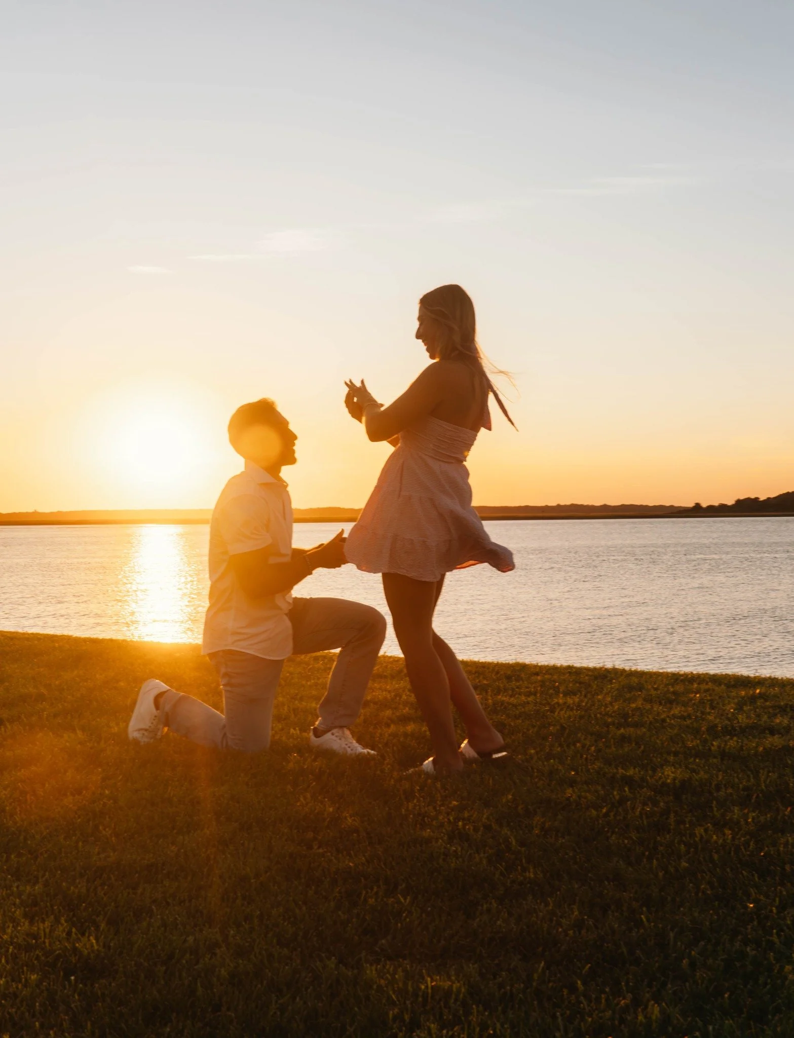 A couple during sunset by a lake, one on one knee proposing, the other smiling with arms raised.