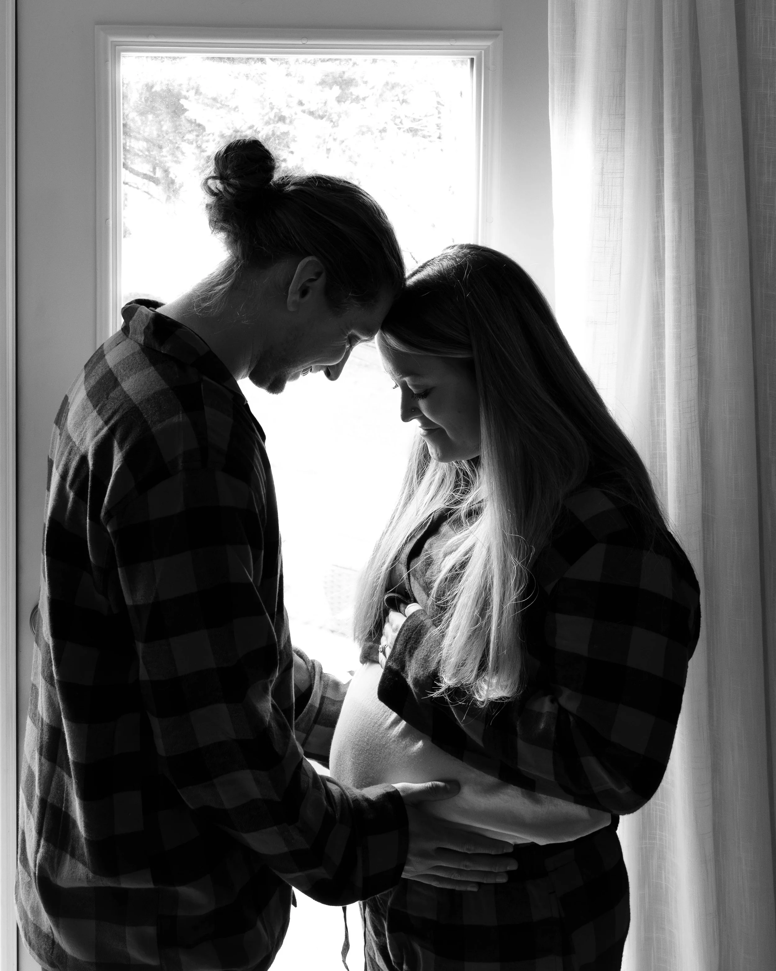 A couple stands in front of a window, with the woman pregnant, touching her belly, and the man resting his hand on her stomach, both smiling and touching foreheads.