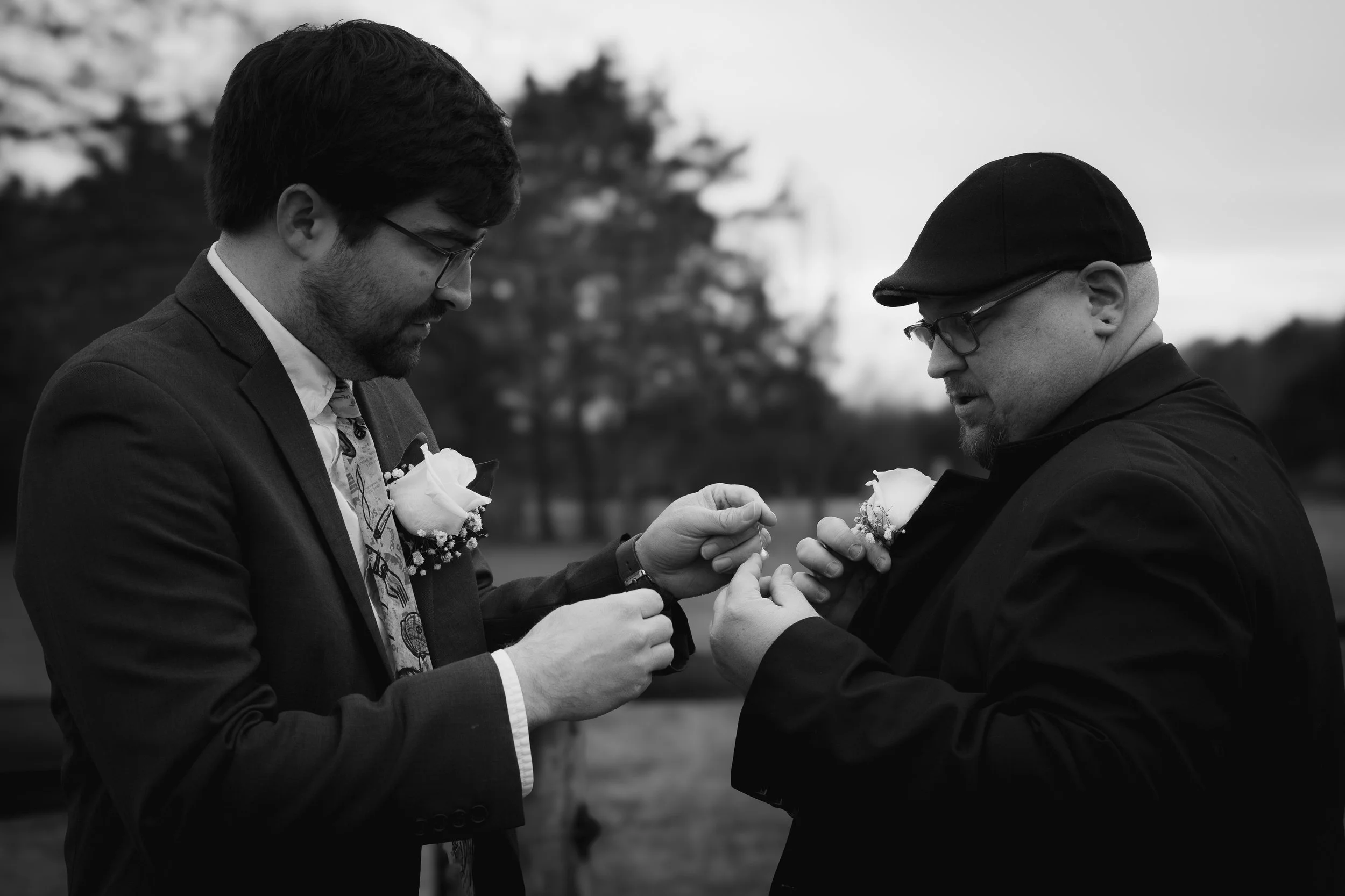 Two men, one younger and one older, dressed in suits with boutonnieres, exchanging rings outdoors in a black-and-white photo, with trees in the background.