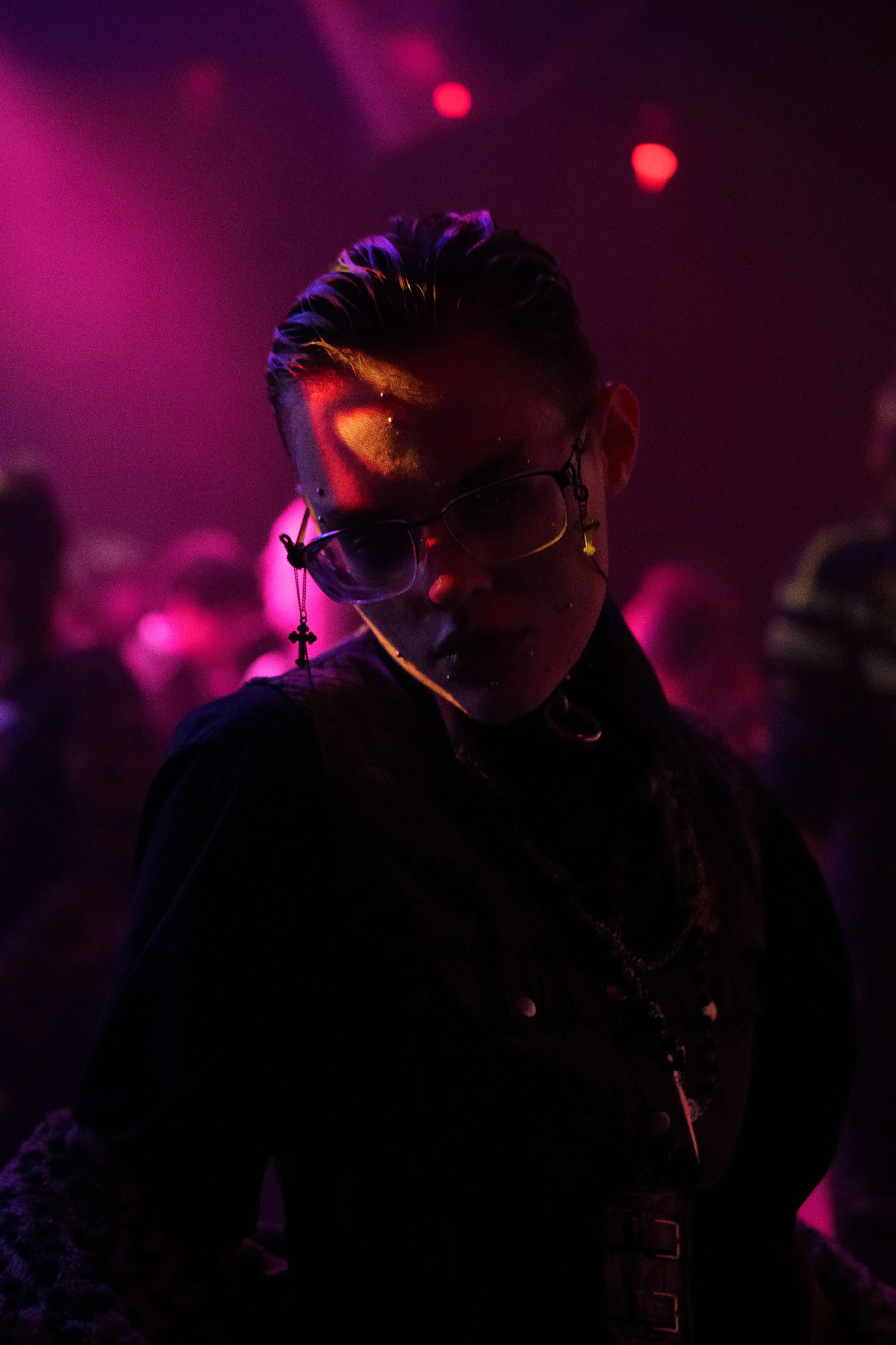 A person with short, styled hair and multiple facial piercings wearing glasses with chains in a dark, neon-lit environment with pink and purple lighting, surrounded by a blurred crowd.