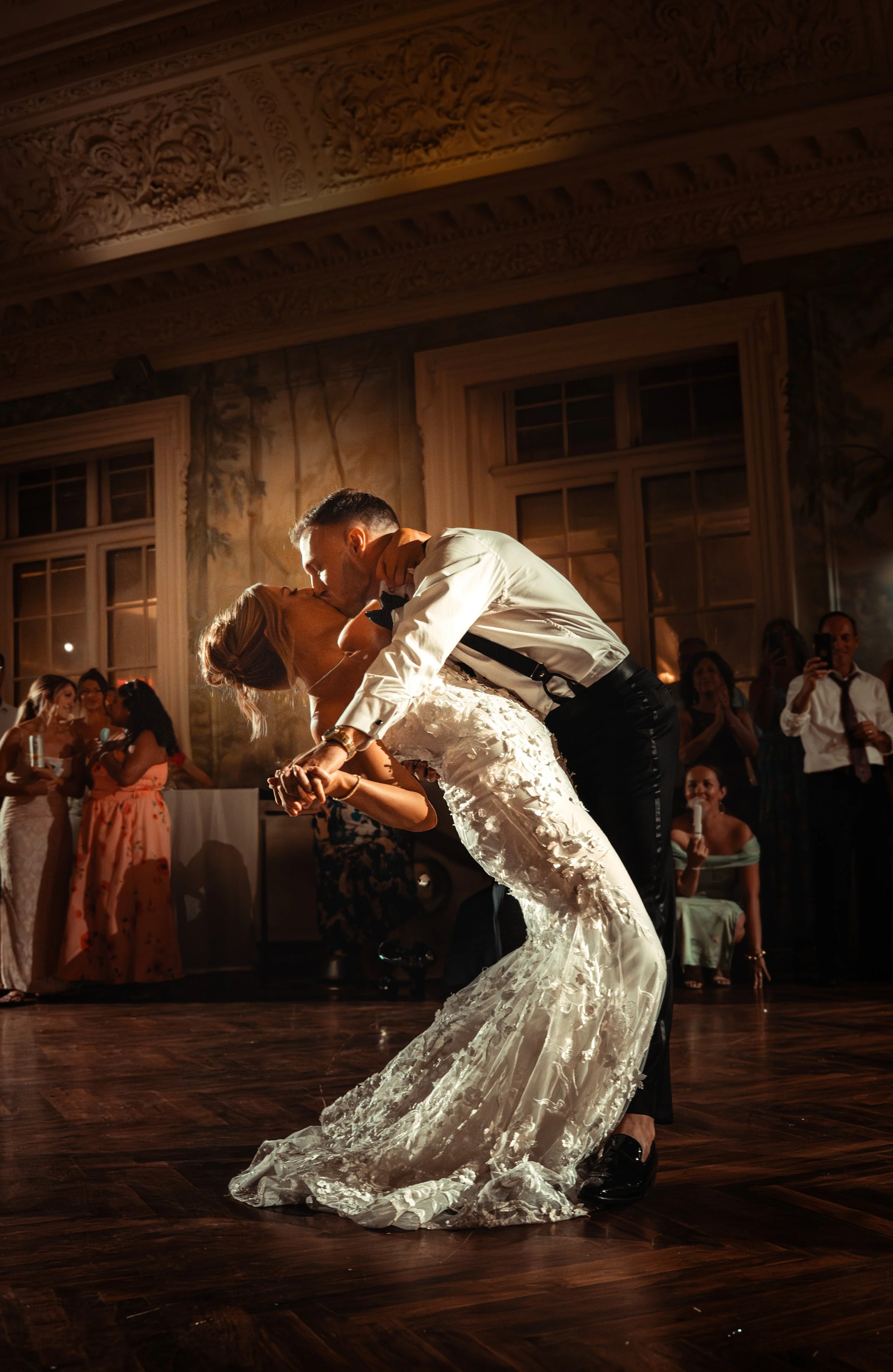 A bride and groom are dancing closely at their wedding reception, surrounded by guests who are watching and taking photos in an elegant room with large windows and ornate decorations.