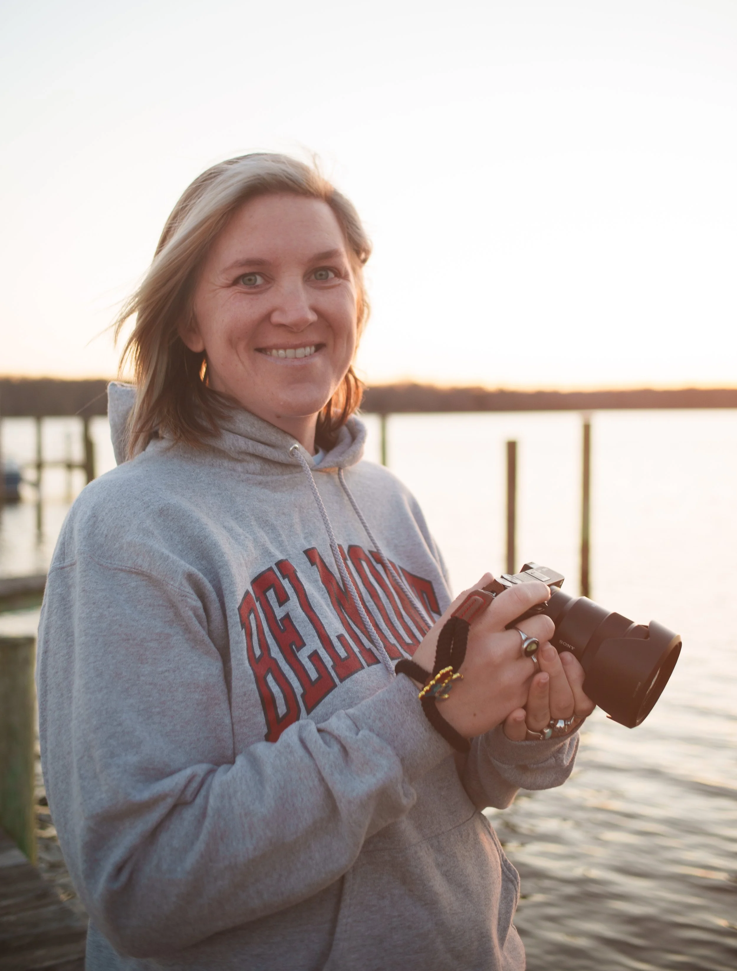 A woman smiling outdoors at sunset, holding a camera, on a wooden dock next to a body of water.