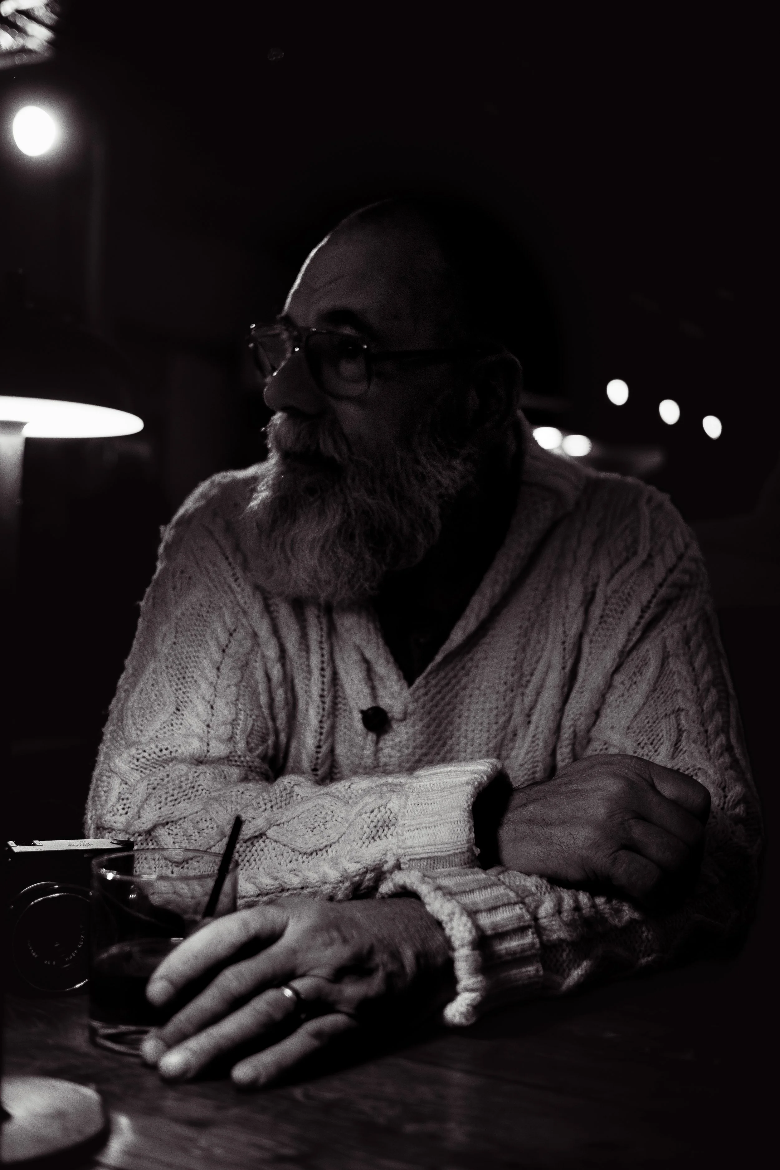 A bearded man with glasses wearing a cable-knit sweater sitting at a dark wooden table in a dimly lit setting, with a glass of drink and a camera on the table.