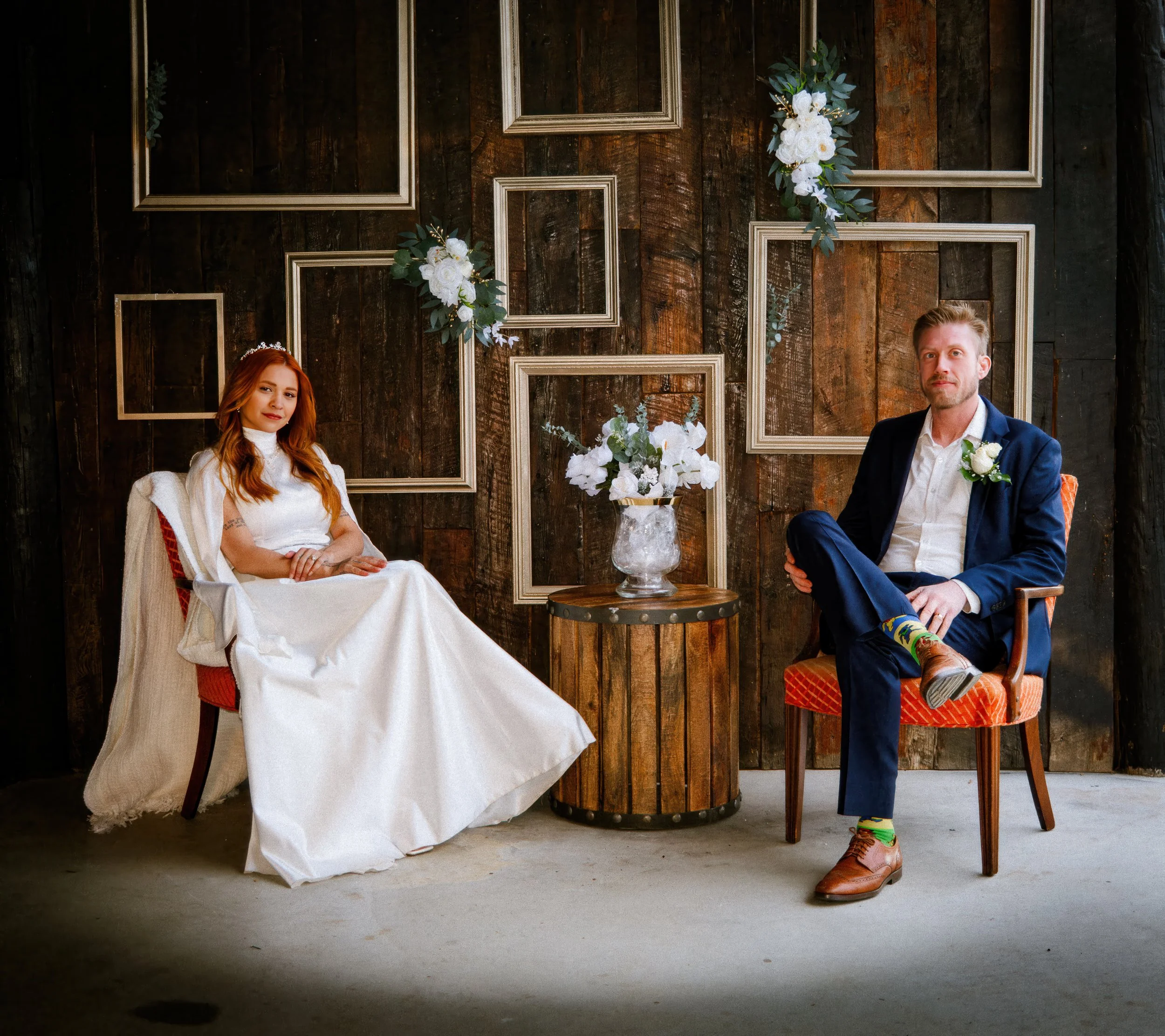 A woman in a white wedding dress and a man in a navy suit with colorful socks sitting on orange chairs in a rustic interior with a wooden wall, empty picture frames, and floral decorations.