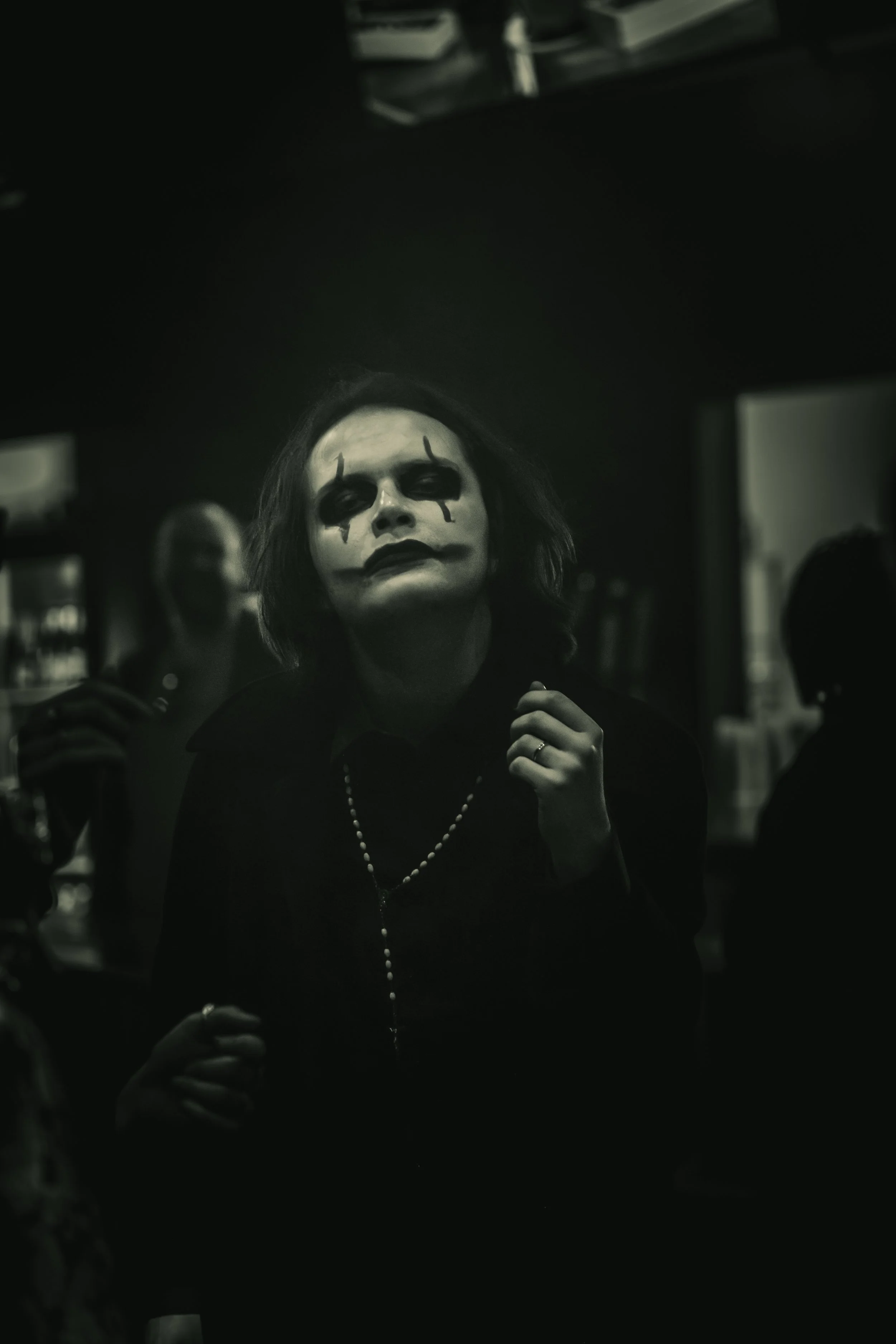 Person with clown makeup, dark clothing, and a pearl necklace, in a dark, dimly lit room.