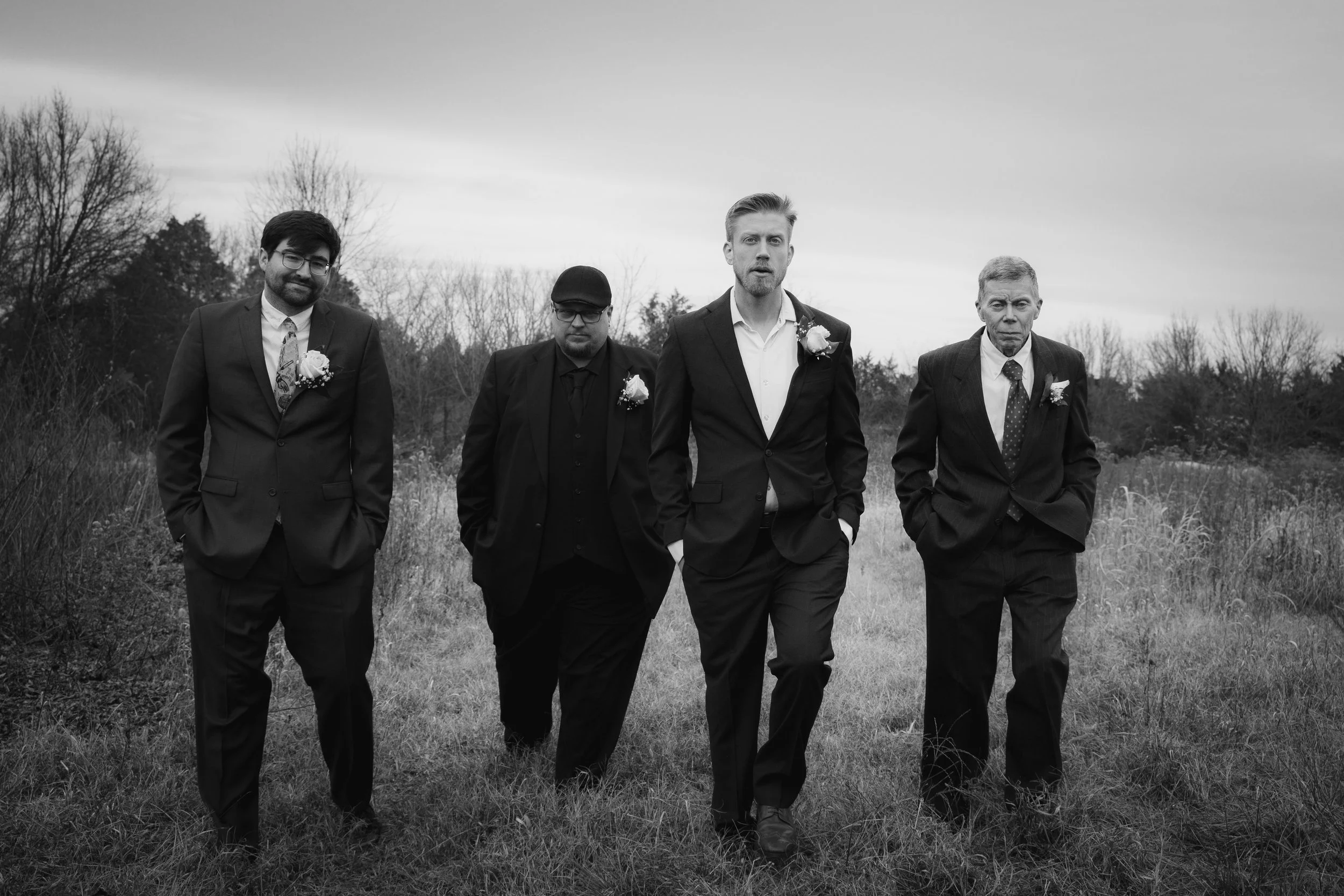 Four men walking outdoors in a field, dressed in suits with boutonnieres, with trees and overcast sky in the background.