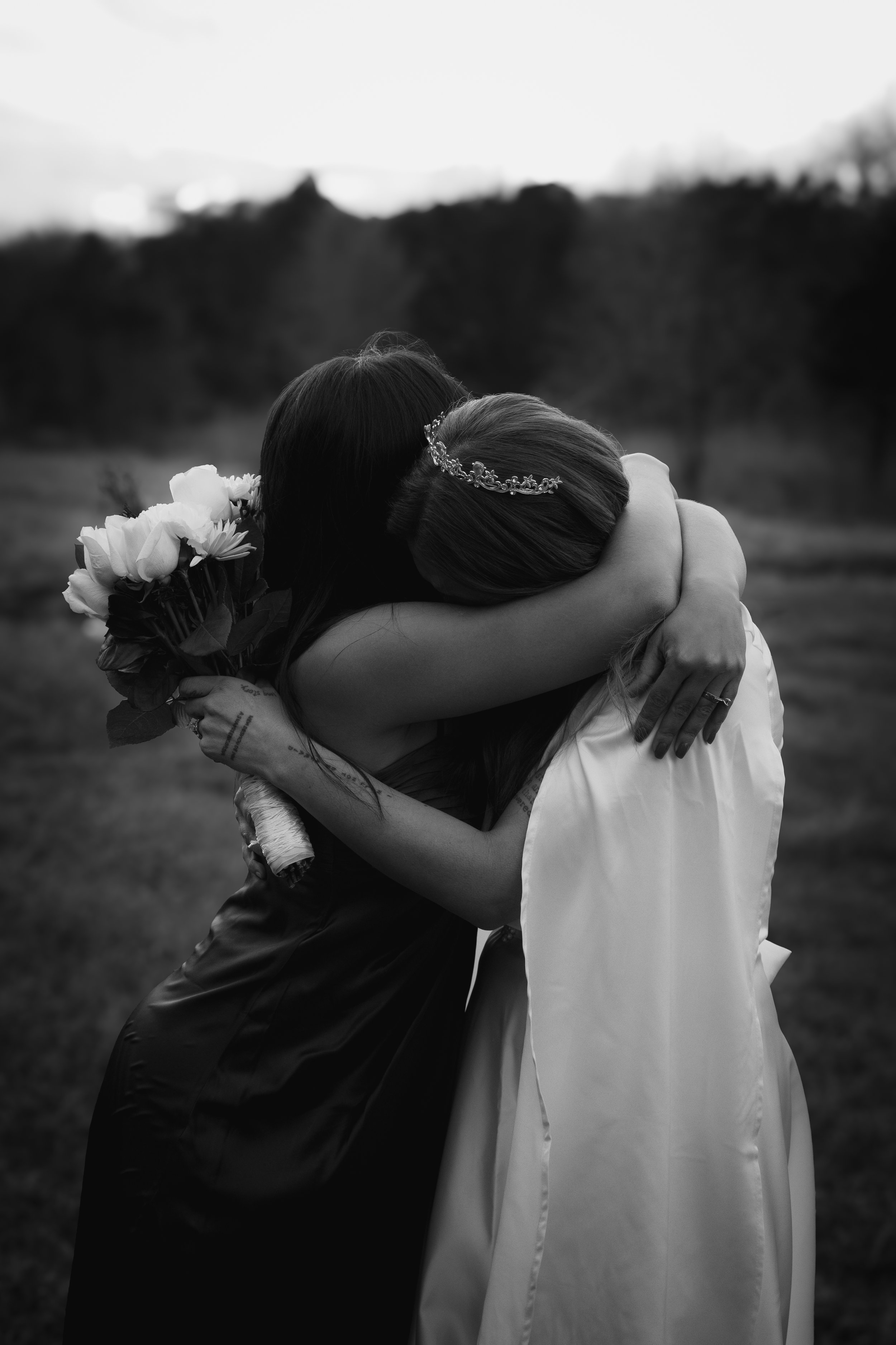 Two women hugging each other outdoors, one in a wedding dress and a tiara, the other holding a bouquet of flowers.