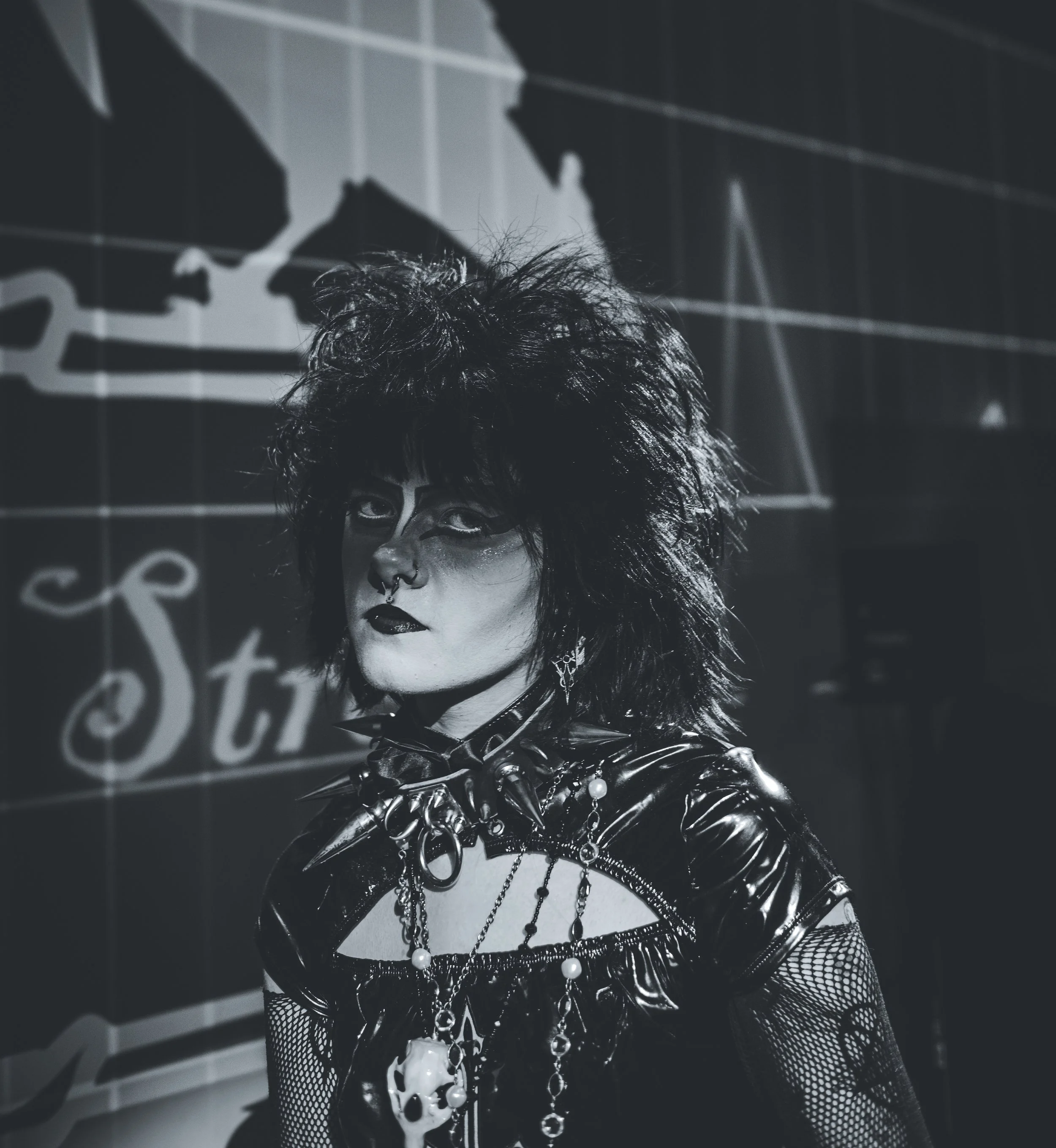 A person with dyed black hair styled in a messy, spiky fashion, wearing dark makeup, a septum piercing, and gothic fashion including a leather jacket adorned with chains and accessories. The background shows a large world map with world names and lin