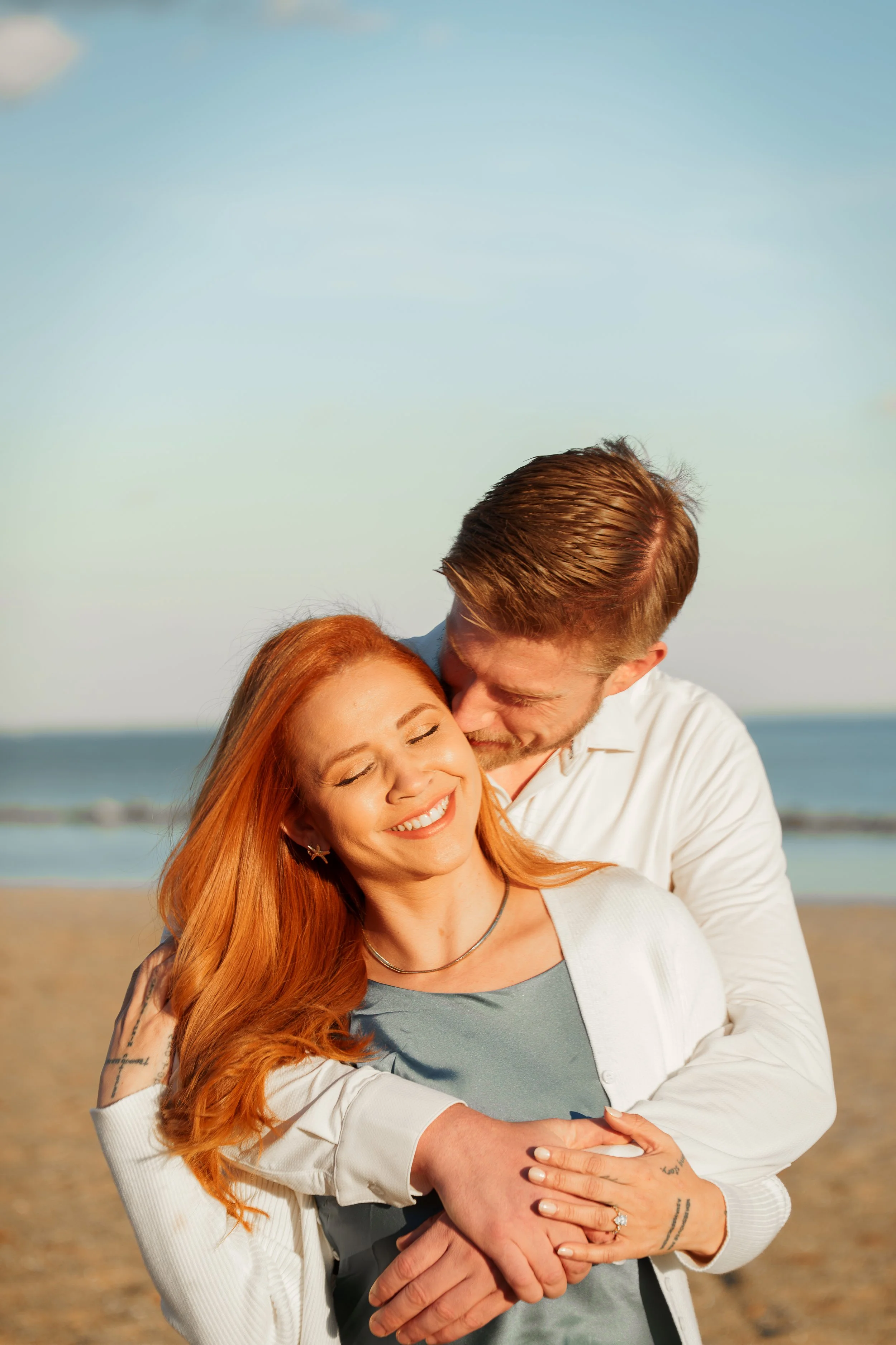 A couple embracing and smiling on a beach with ocean waves in the background.