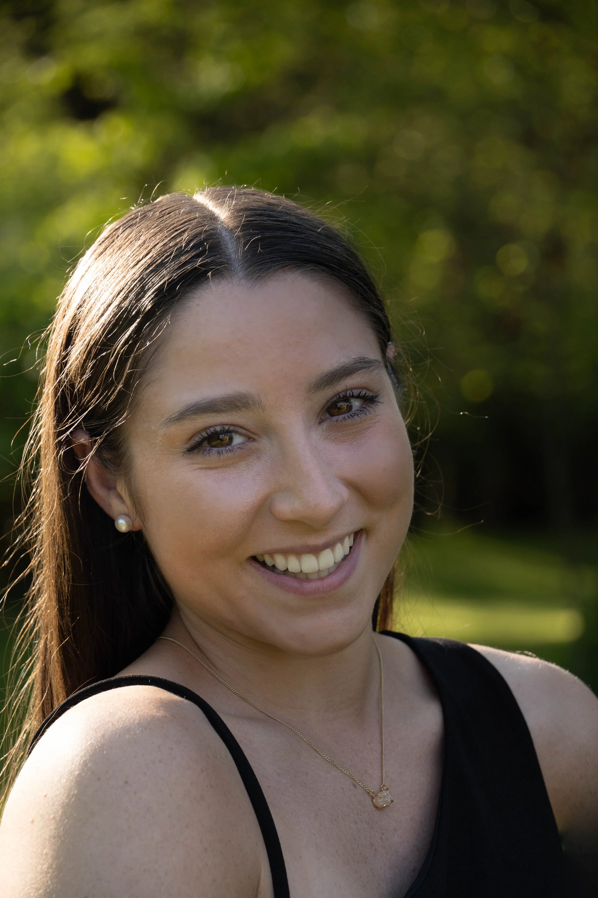 Smiling woman with brown hair wearing pearl earrings and a gold necklace outdoors on a sunny day.