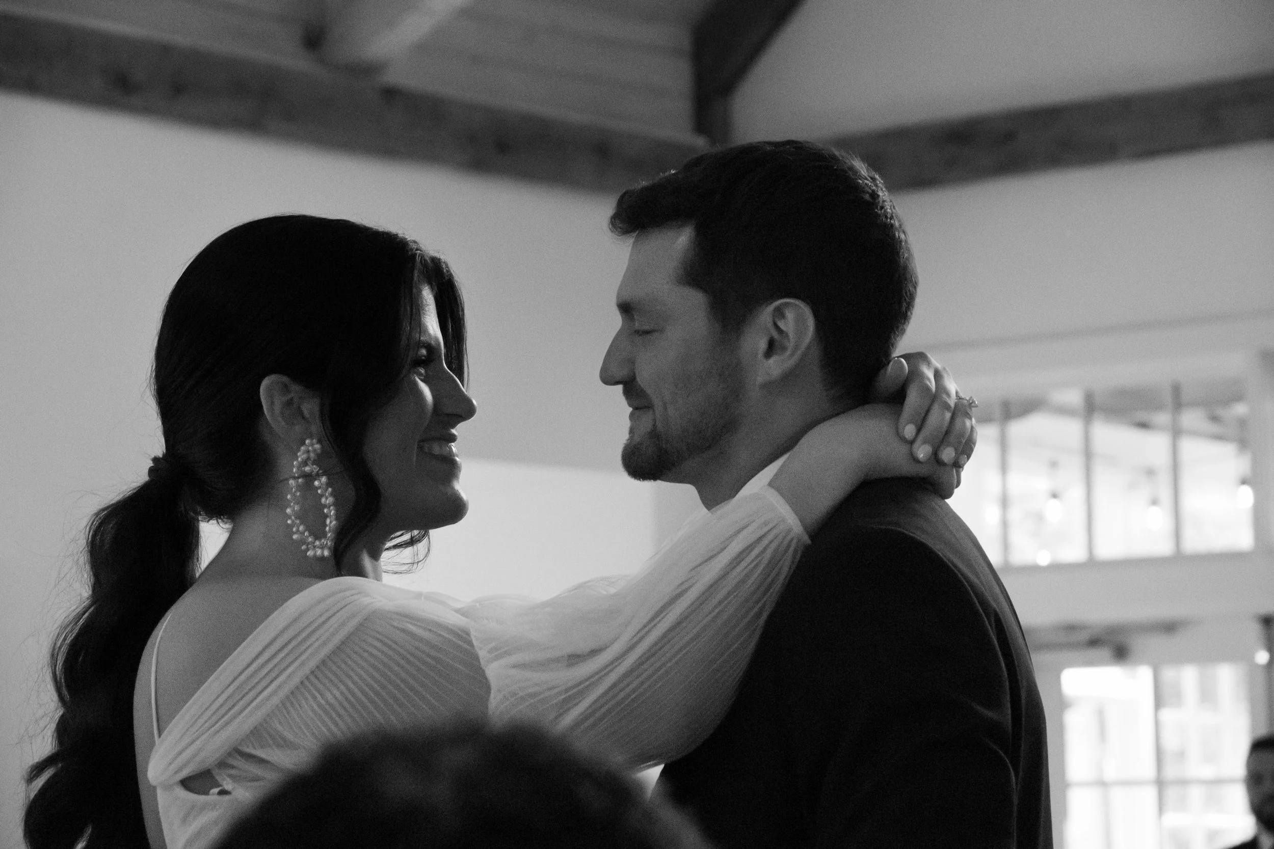 A black and white photo of a smiling woman with long dark hair in a ponytail and large earrings, holding a man by the neck while dancing. The man has short dark hair and a beard, and is dressed in a suit. They are indoors with large windows in the ba