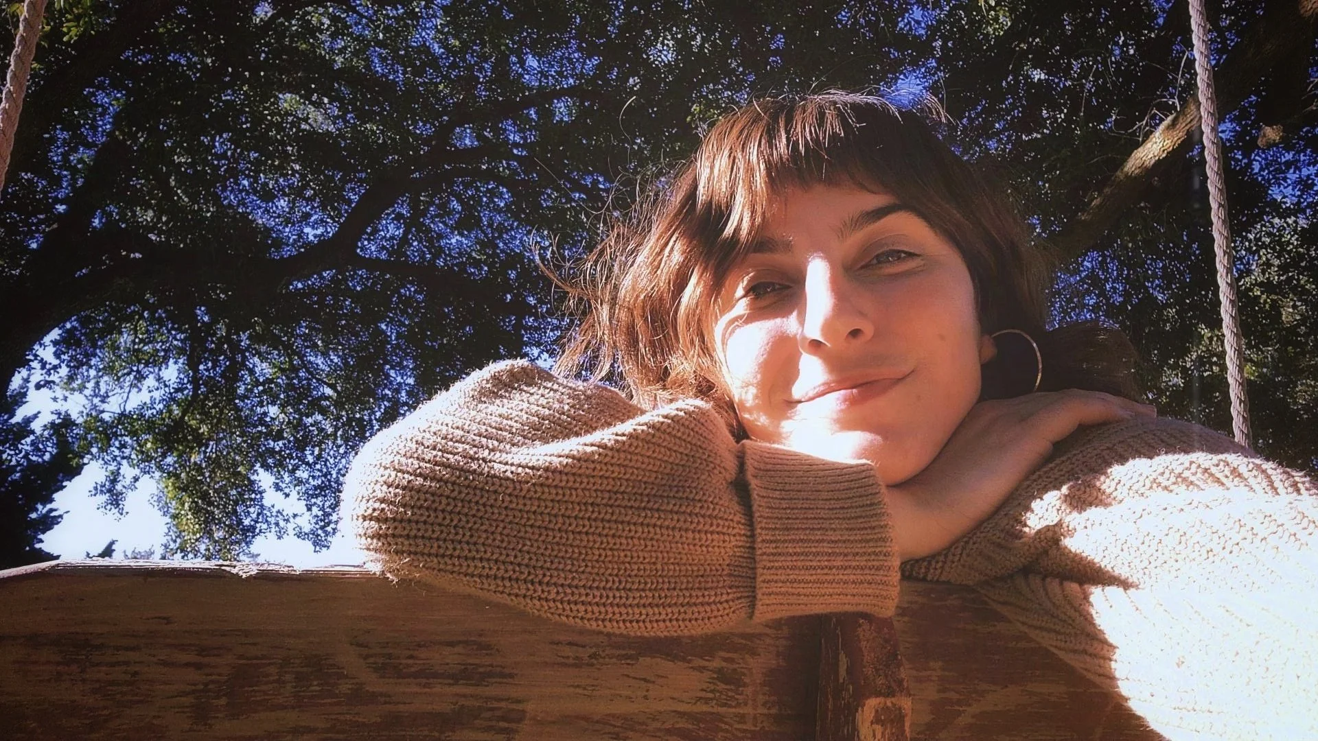 Young woman with brown hair and hoop earrings resting her chin on her hands, smiling softly while sitting outdoors under a large tree with green leaves and sunlight filtering through.