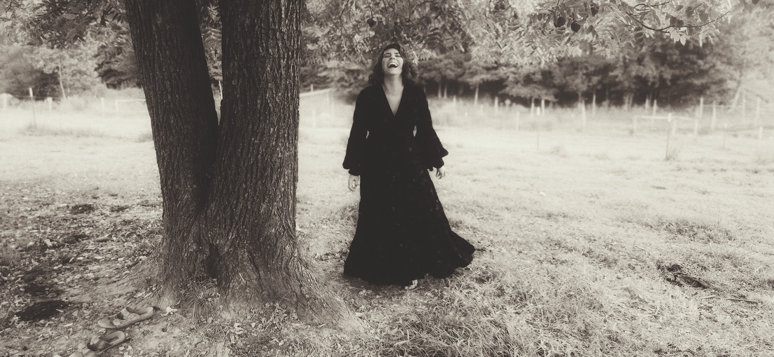 A woman in a long black dress laughing and standing outdoors near a large tree, in a grassy area with a fence in the background, in black and white.