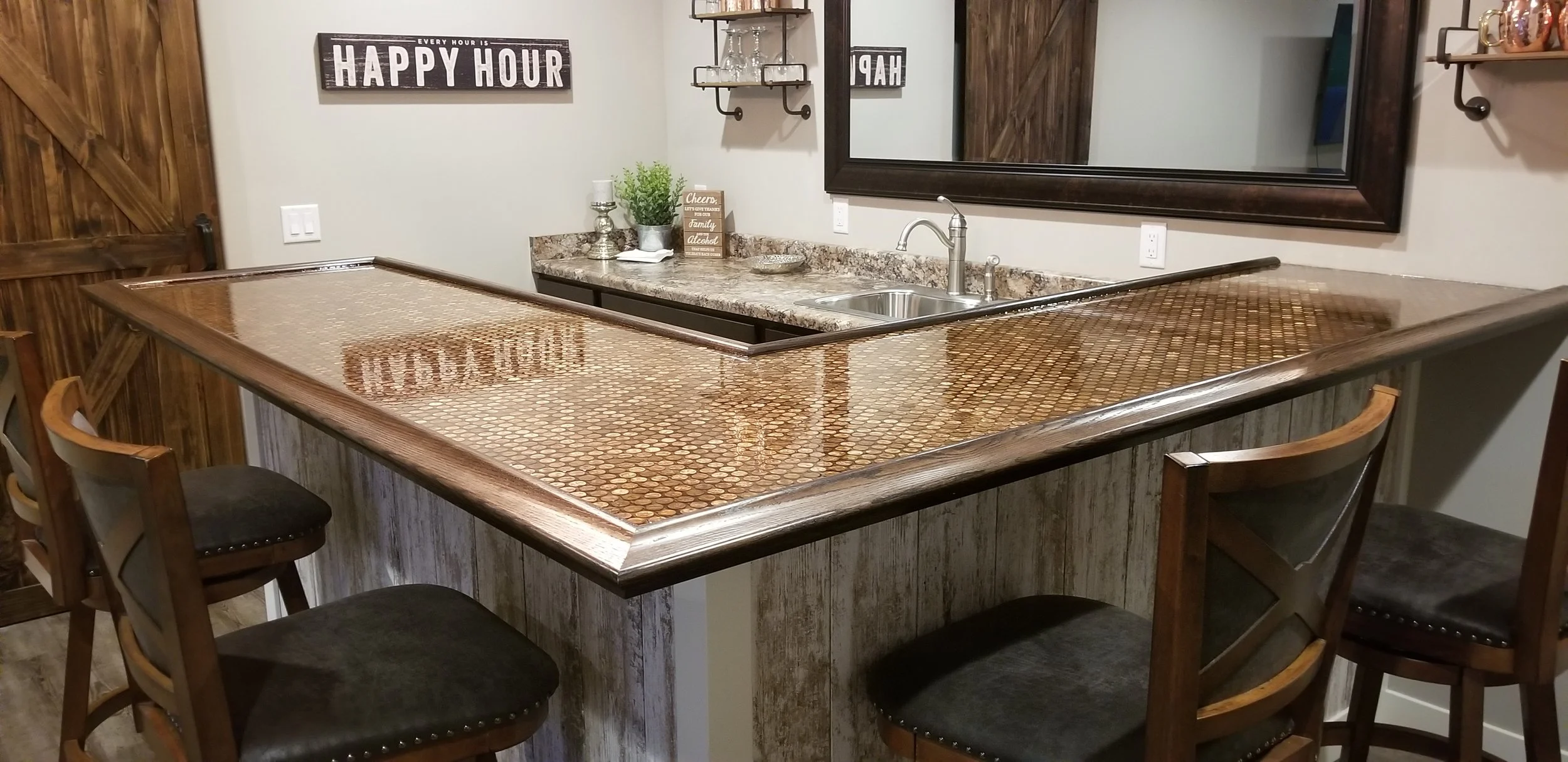 A kitchen island with a copper penny tile countertop, a small sink, and wooden chairs around it. Behind the island, there is a granite countertop with a small plant, a sign, and decorations, a large mirror on the wall, and a barn door.