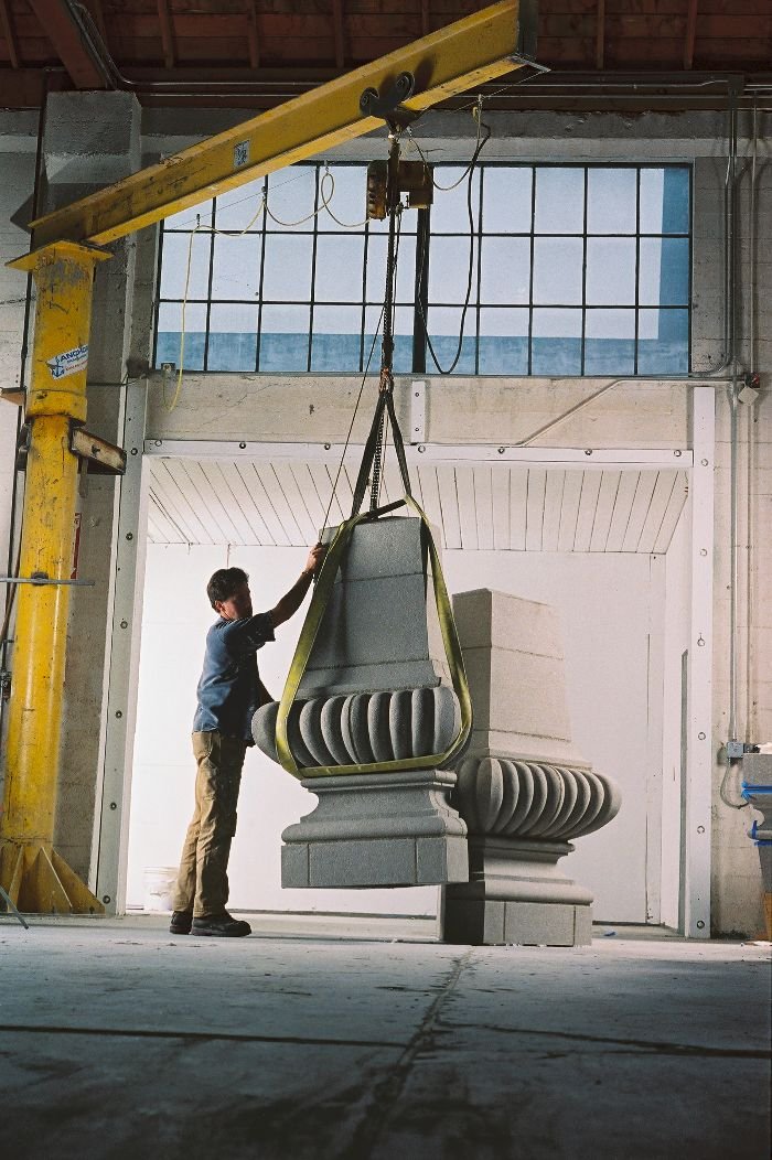 A worker in a warehouse or workshop is guiding a large stone or concrete sculpture with a hoist, lifting it to an upright position.
