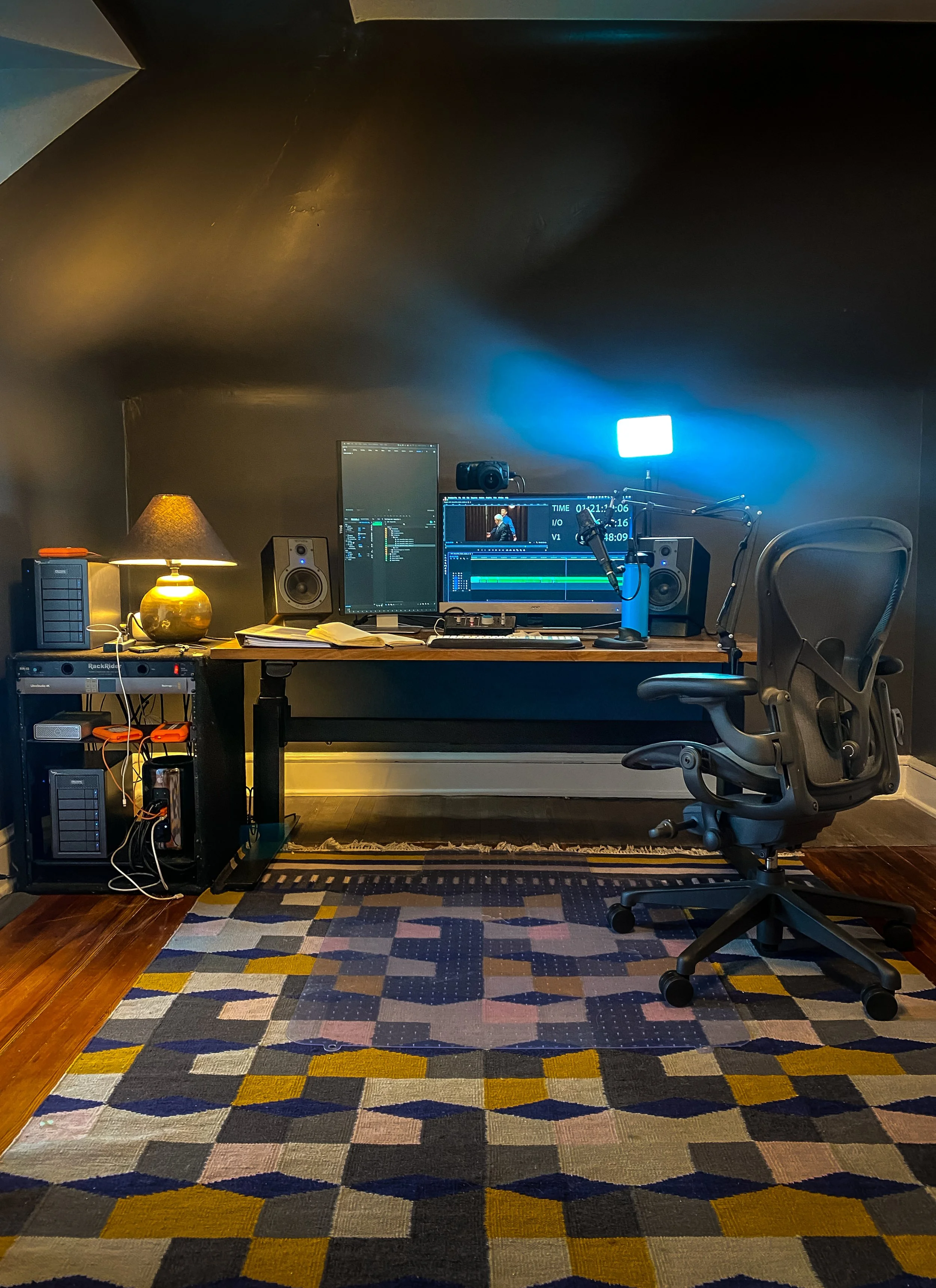A home office setup with a desk, two monitors, a microphone, and speakers. There is a wooden desk lamp on a side unit, a chair, and a colorful patterned rug on a wooden floor.