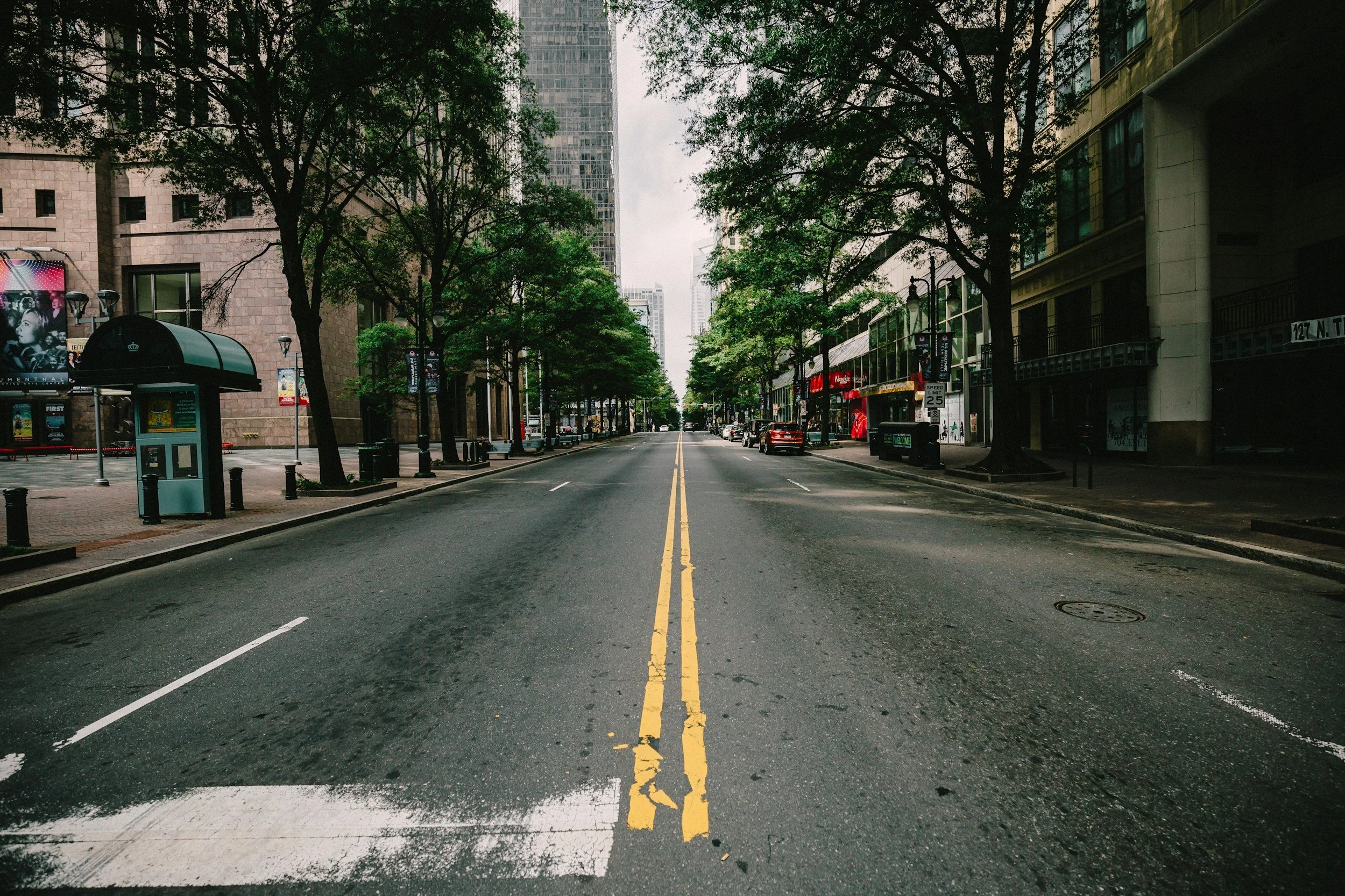 Empty city street with trees, buildings, and parked cars on both sides, under cloudy sky.