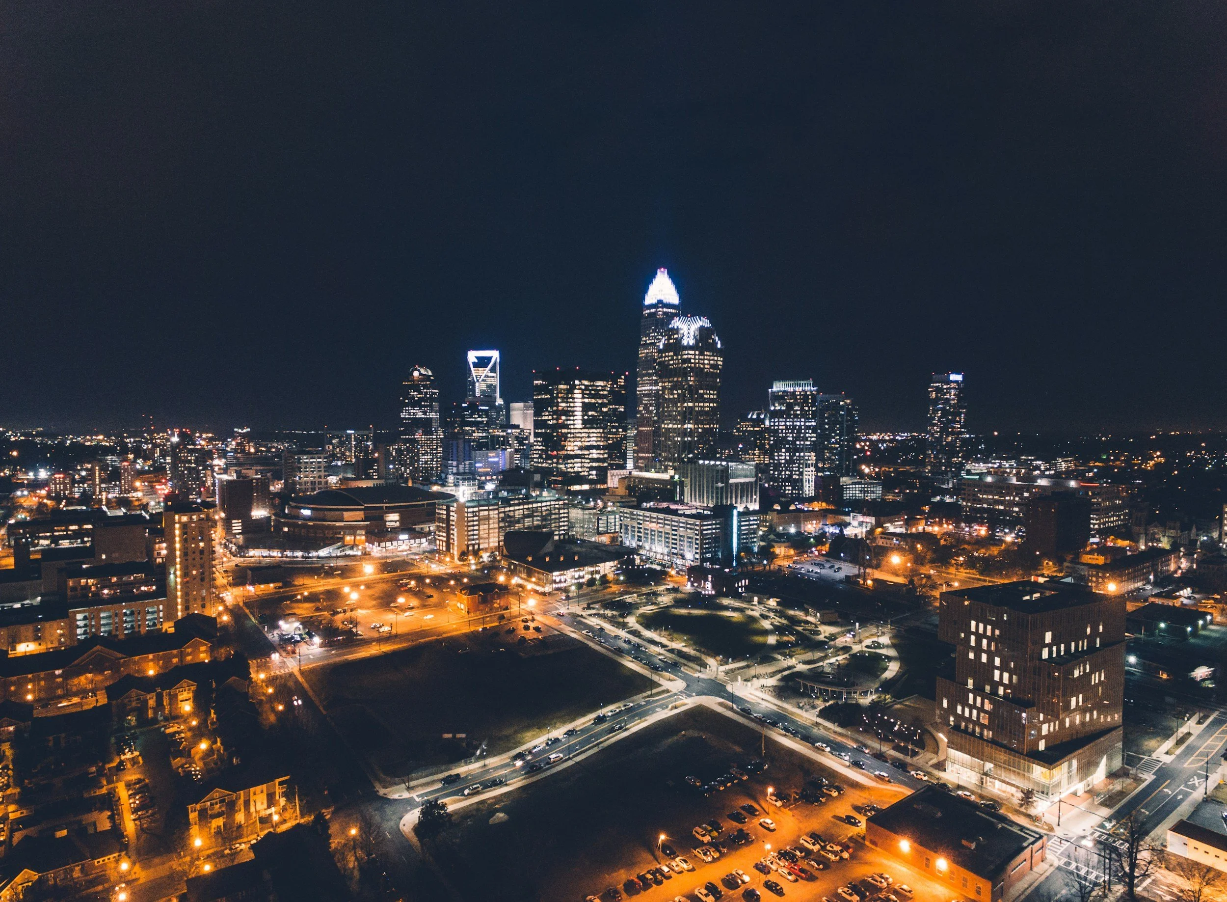 Nighttime cityscape of a downtown area with illuminated skyscrapers and surrounding buildings, parking lot, and streets with cars.