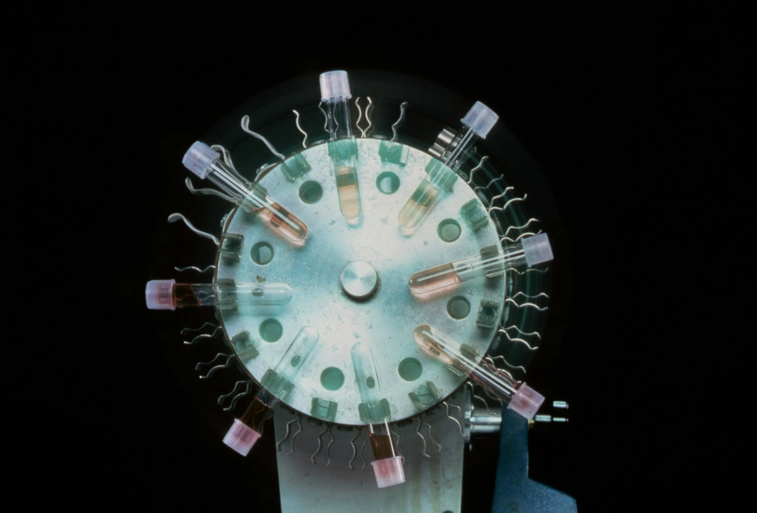 Microscope stage with multiple glass test tubes filled with different colored liquids arranged in a circular pattern, set against a dark background.