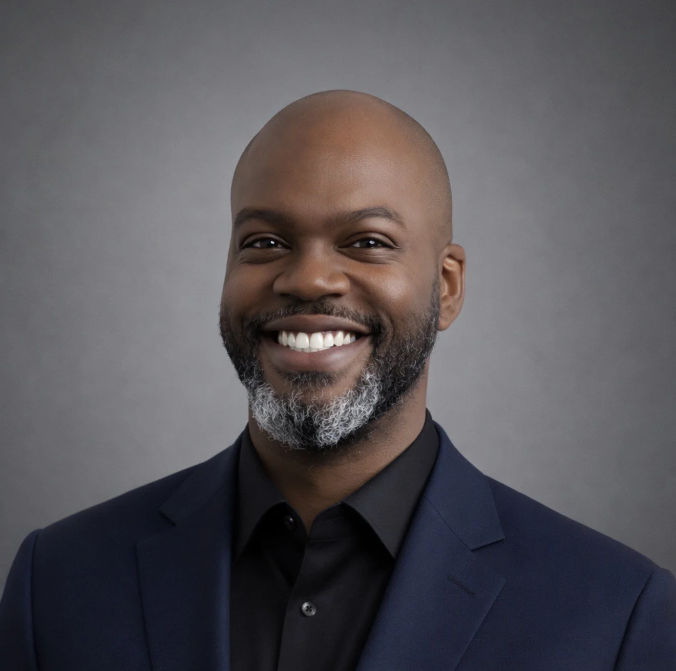 A smiling man with a beard and mustache, wearing a dark suit and black shirt, against a gray background.