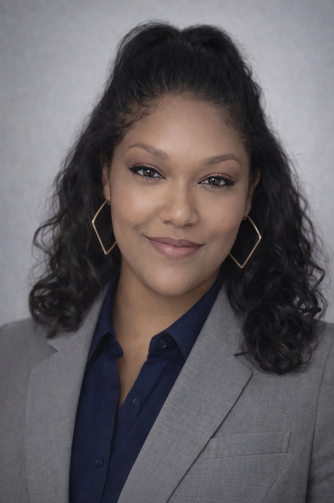 A professional woman with dark, curly hair worn in loose ringlets, wearing a gray blazer, dark blue shirt, gold geometric earrings, and smiling against a gray background.