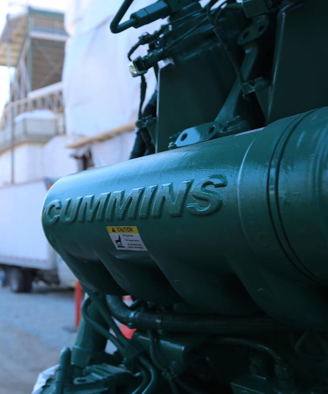 Close-up of a green Cummins industrial engine with a caution sticker, outdoors setting with construction or industrial background.