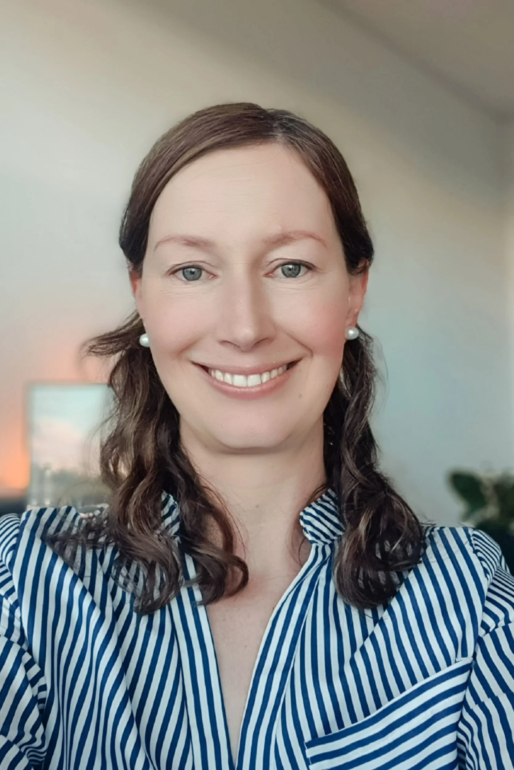 A woman with shoulder-length curly brown hair, wearing pearl earrings and a blue and white striped blouse, smiling indoors with a blurred background.