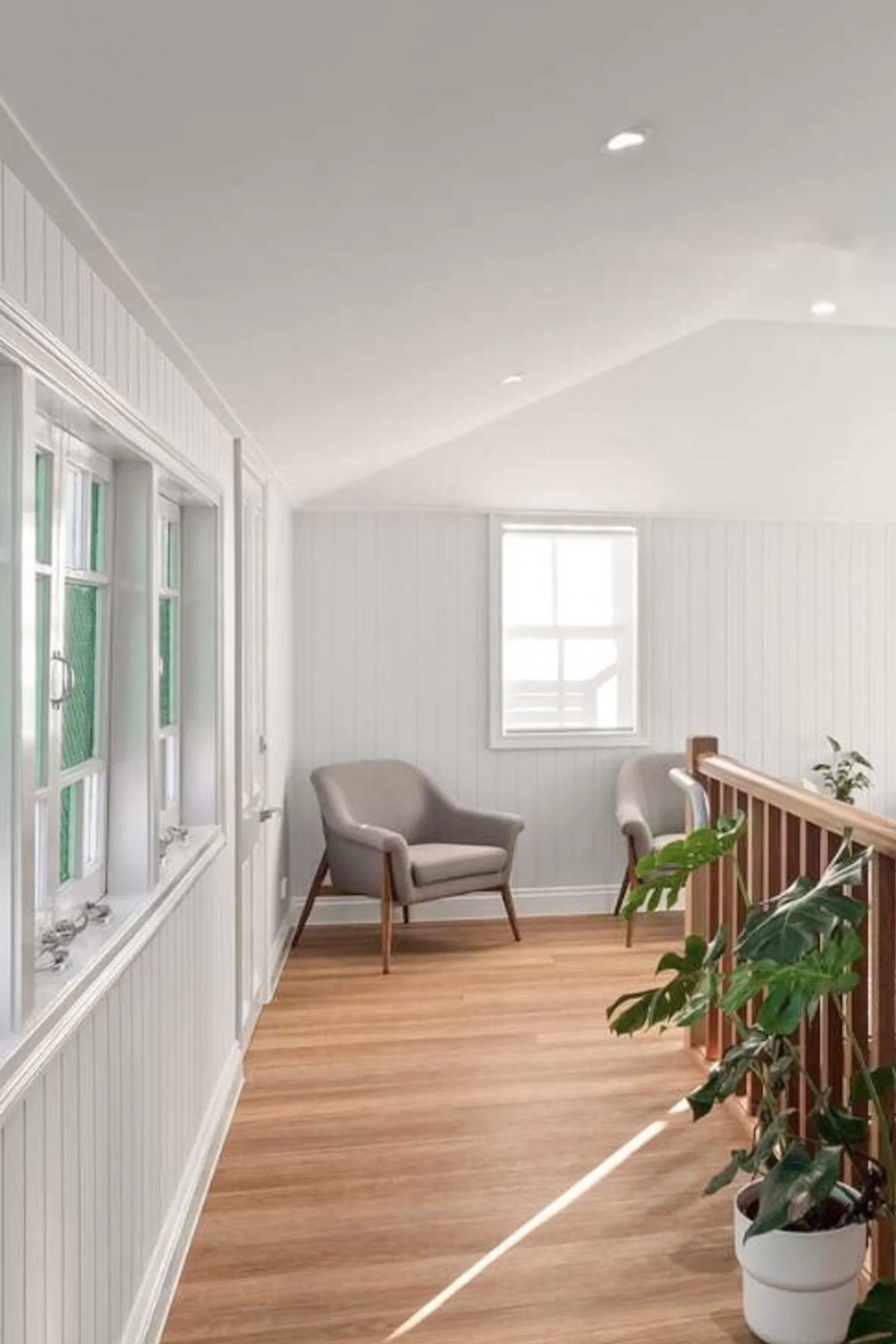 Bright upstairs room with white paneled walls, wooden floor, two cream-colored armchairs, a window, and a potted plant on a wooden railing.
