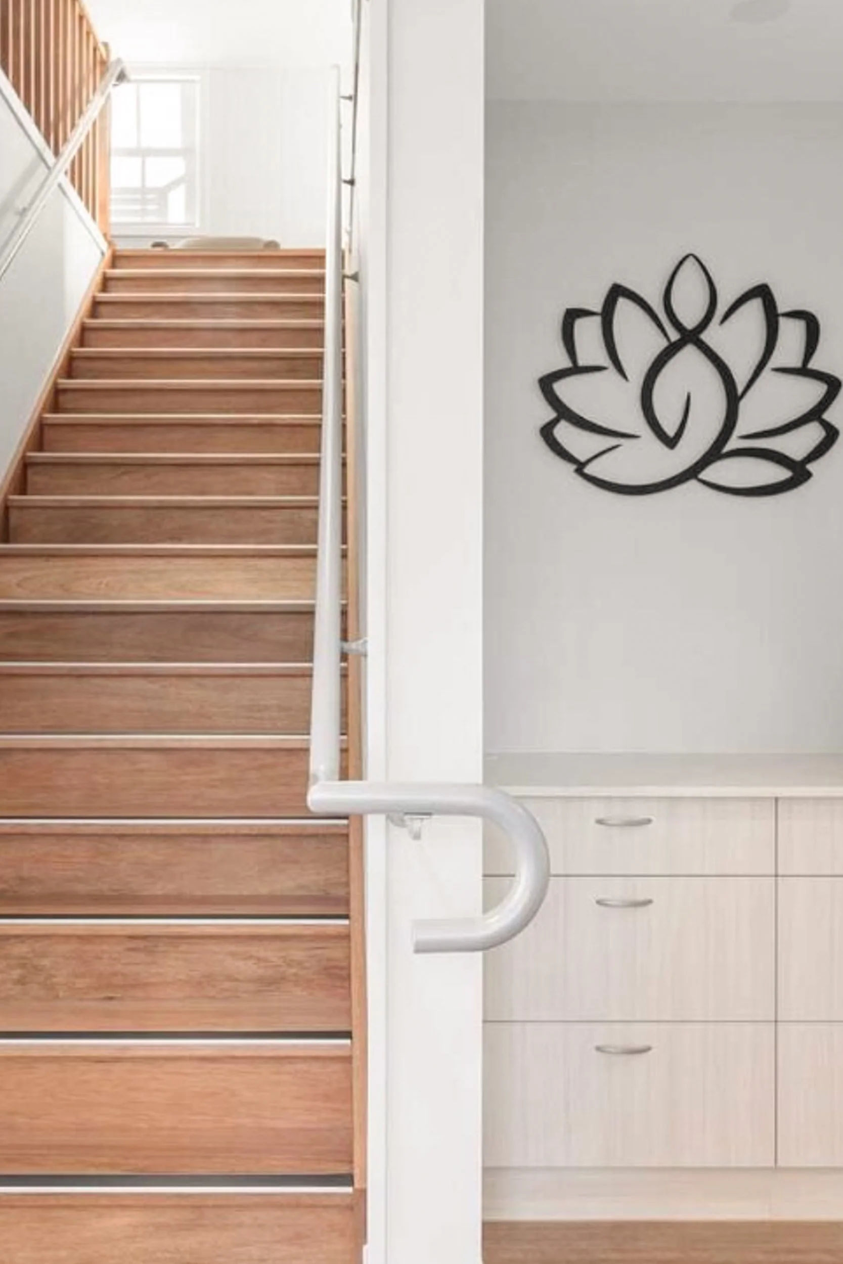 Wooden staircase with metal handrails, white walls, a window at the top, and a black wall decoration resembling a stylized lotus flower or mandala.