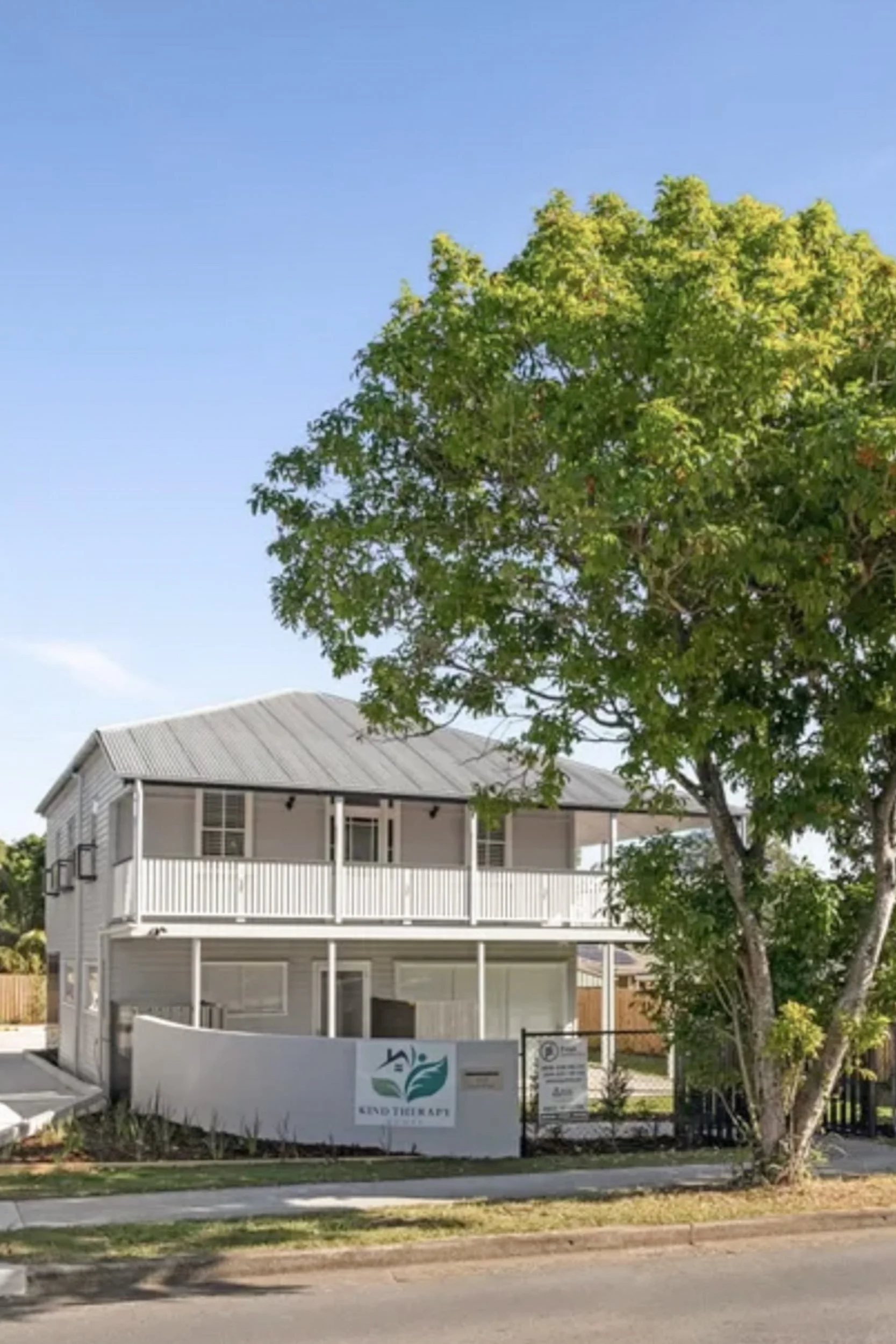 Two-story white and grey Queenslander house with a large green tree in front, screened porch, and a sign for Kind Therapy House with a leaf logo.