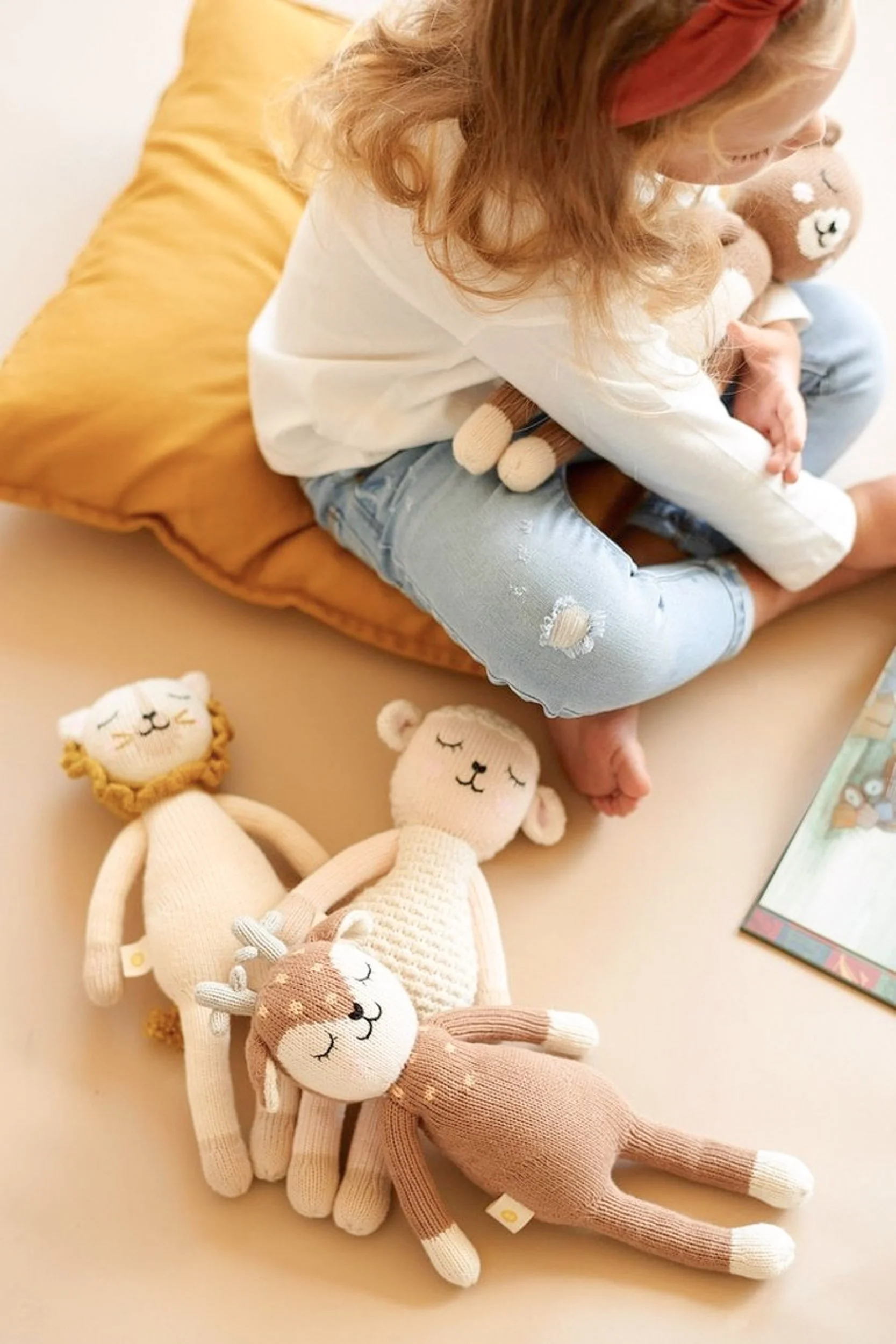 A young girl sitting on a yellow pillow with a teddy bear plush toy on her lap. She is wearing a red headband, white long-sleeve shirt, and ripped jeans. In front of her on the surface are four stuffed animal dolls: a lion, a bear, a rabbit, and a deer, all with closed eyes and smiling faces, made of soft knitted fabric.