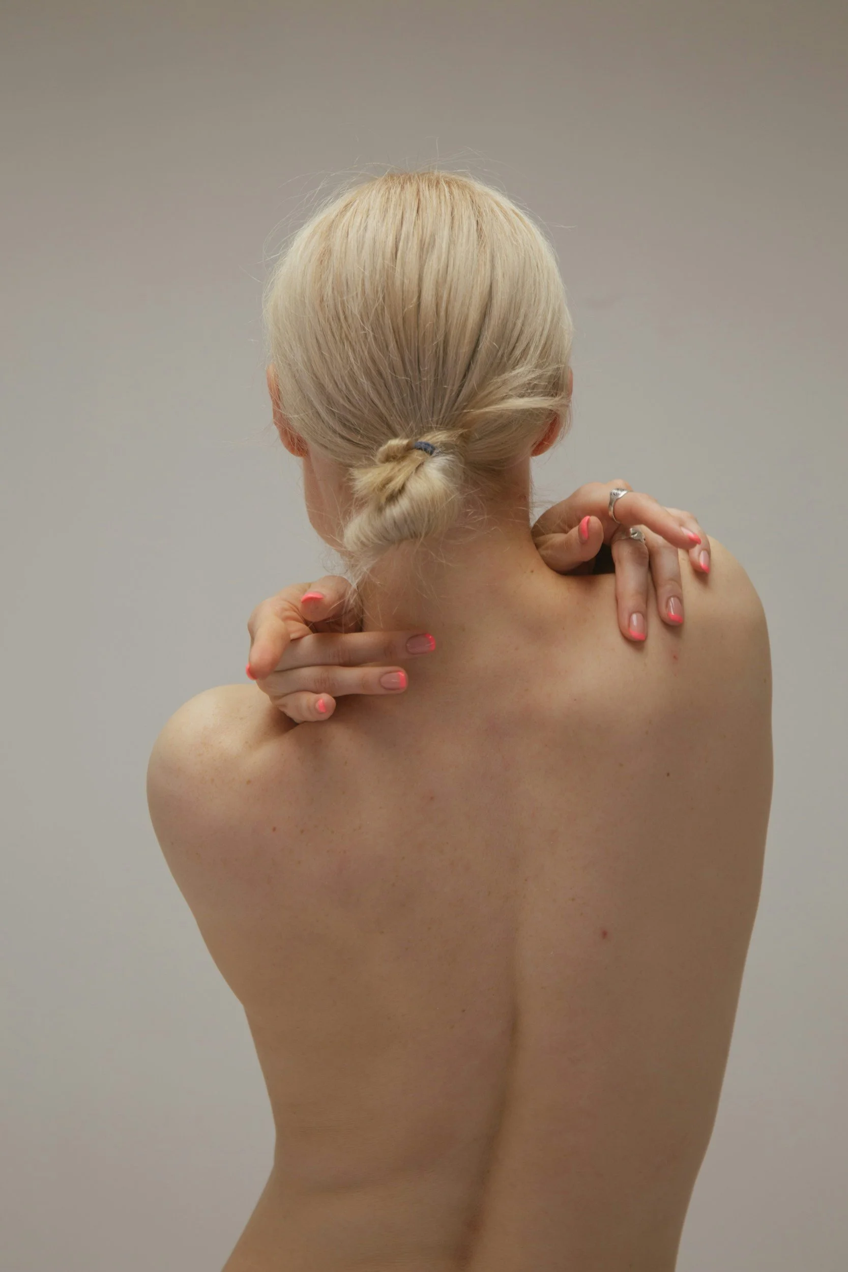 A woman with blonde hair tied in a low bun, facing away, touching her shoulders with both hands, revealing her back and shoulders.