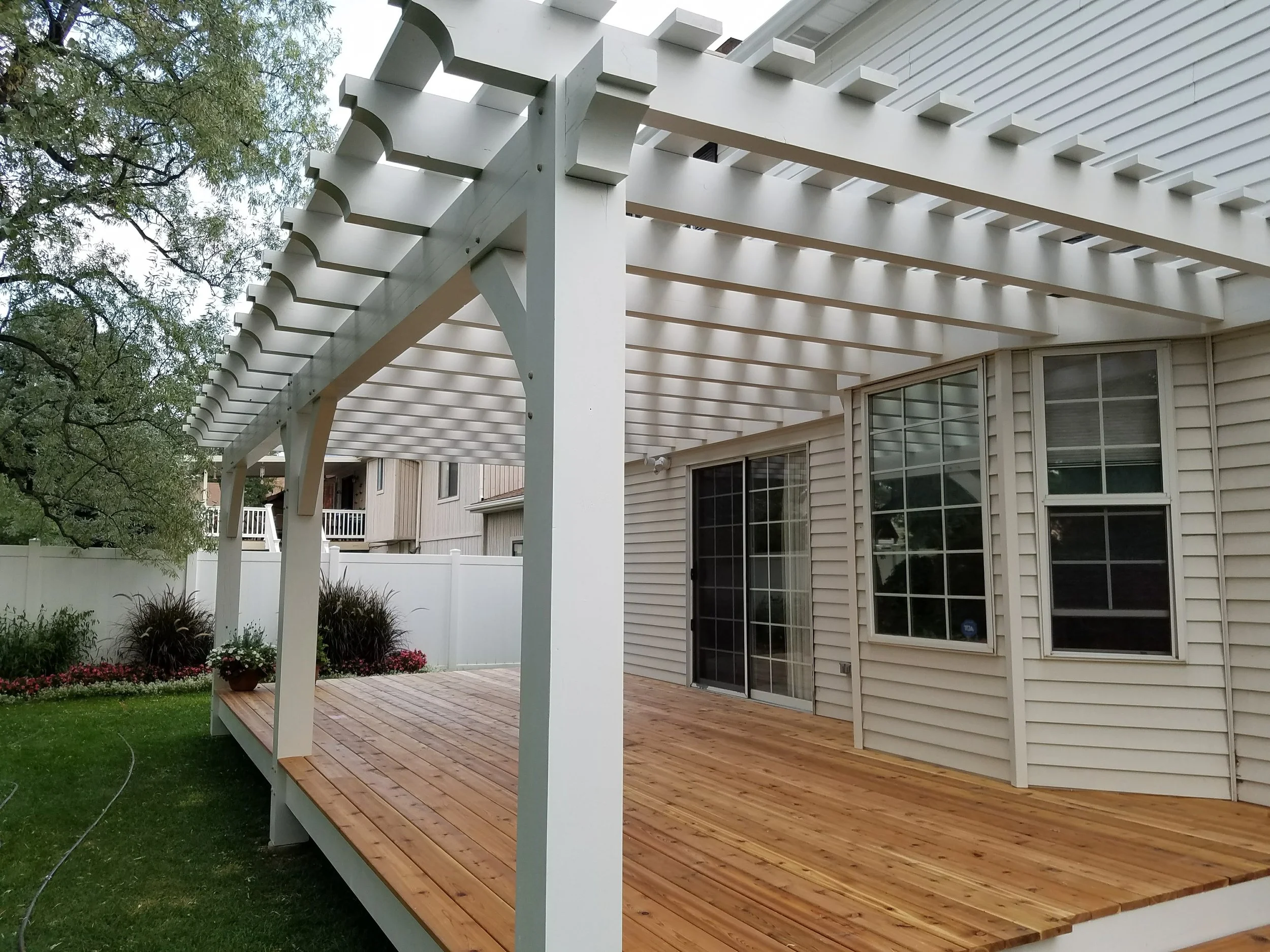Backyard wooden deck with white pergola frame, surrounded by a grassy yard, garden plants, and a white vinyl fence.