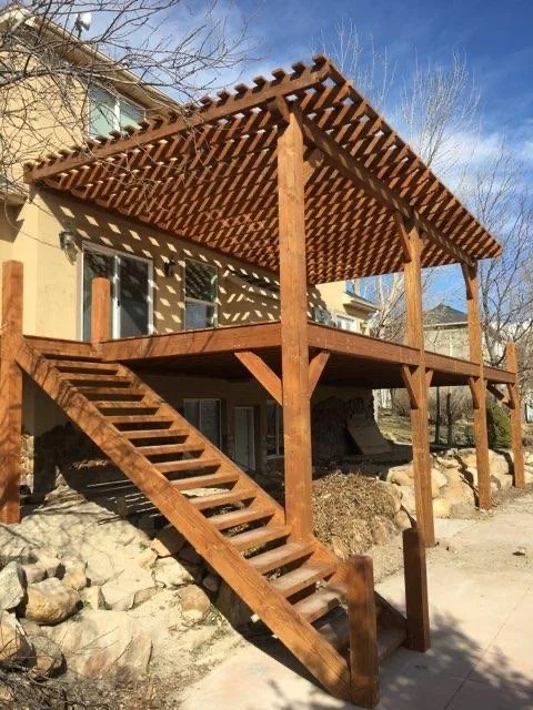 A timber deck with stairs leading up to the second-story balcony pergola, attached to a house. 